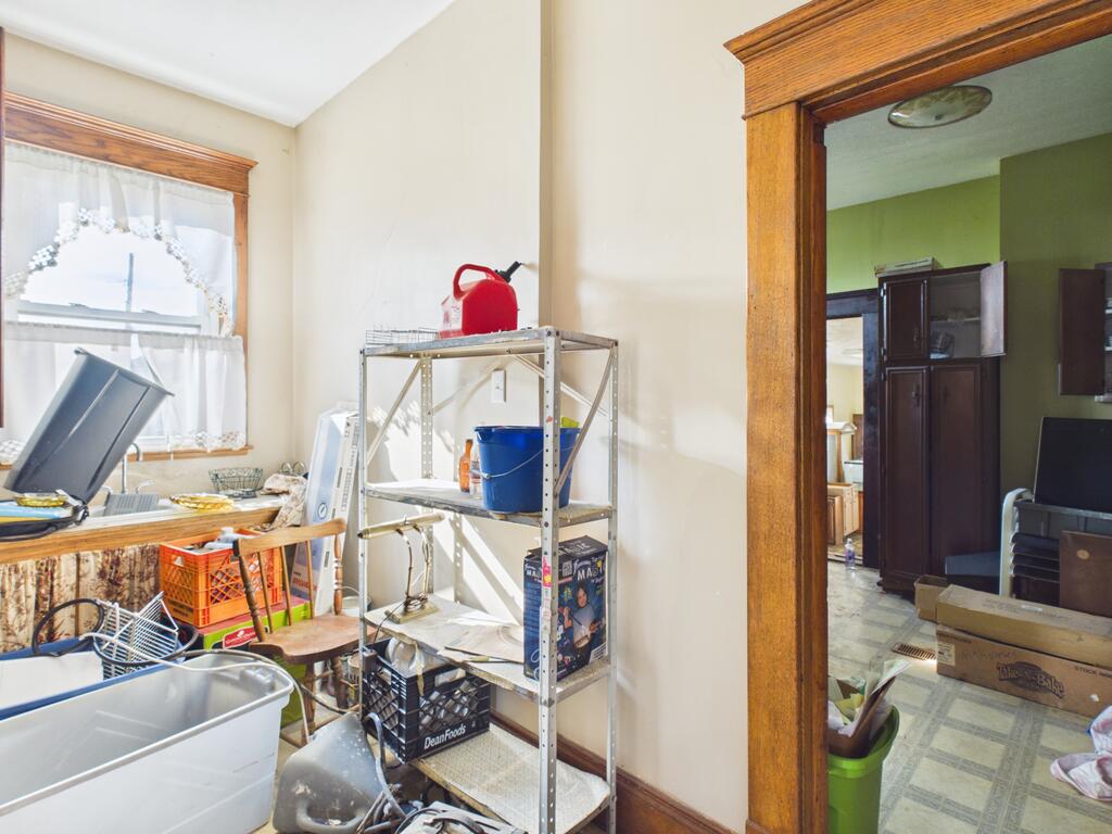 Cluttered utility room with a deep sink, metal shelving, and an open doorway to a green-walled room.