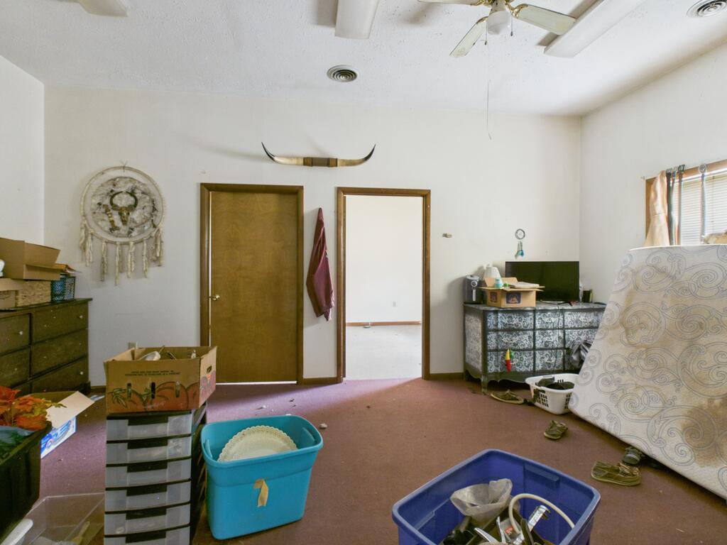 Cluttered bedroom with mauve carpet, storage bins, and a mattress leaning beside the window.