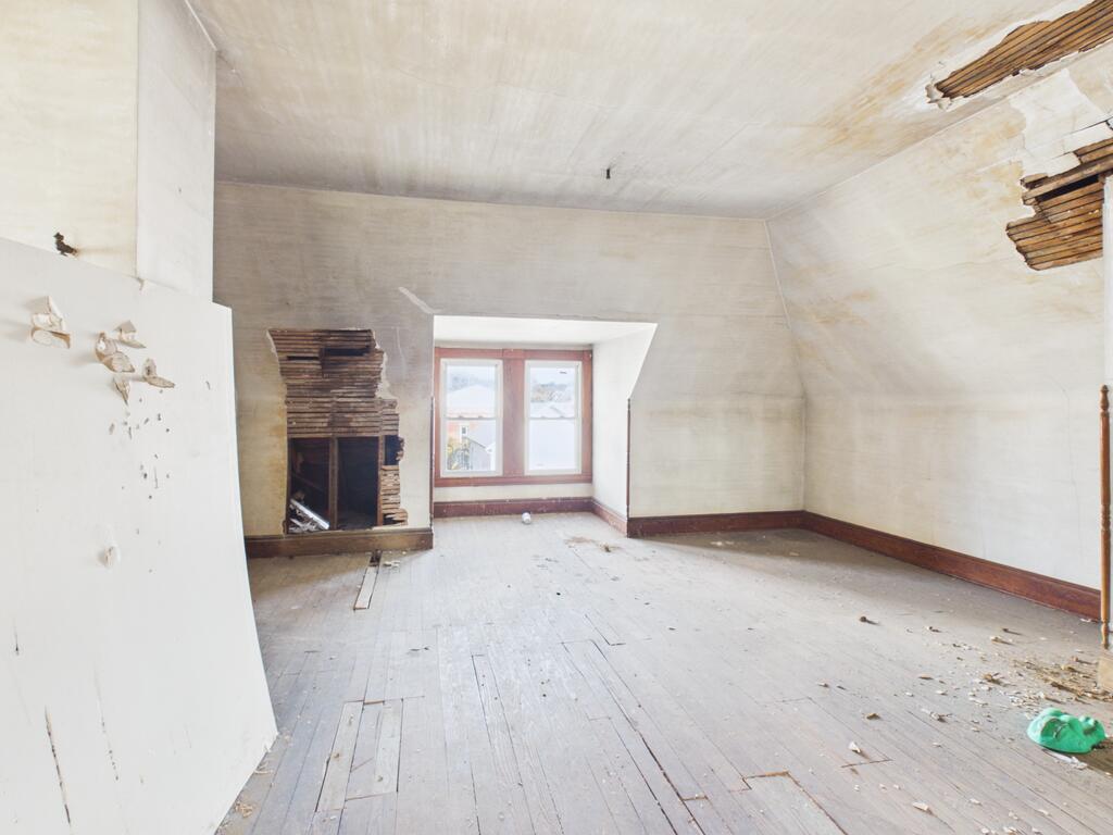 Vacant room with a damaged fireplace, exposed lath, and two far windows.