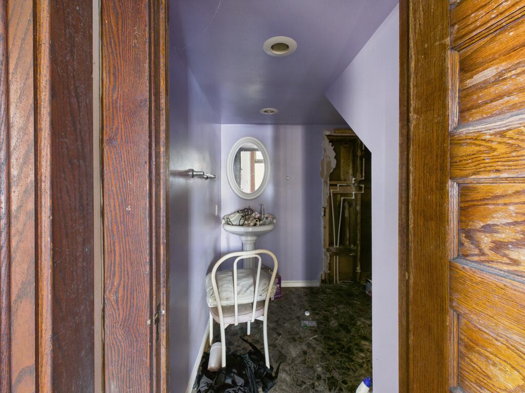 Narrow lavender bathroom with a pedestal sink, oval mirror, ornate wooden doorway, and damaged wall.