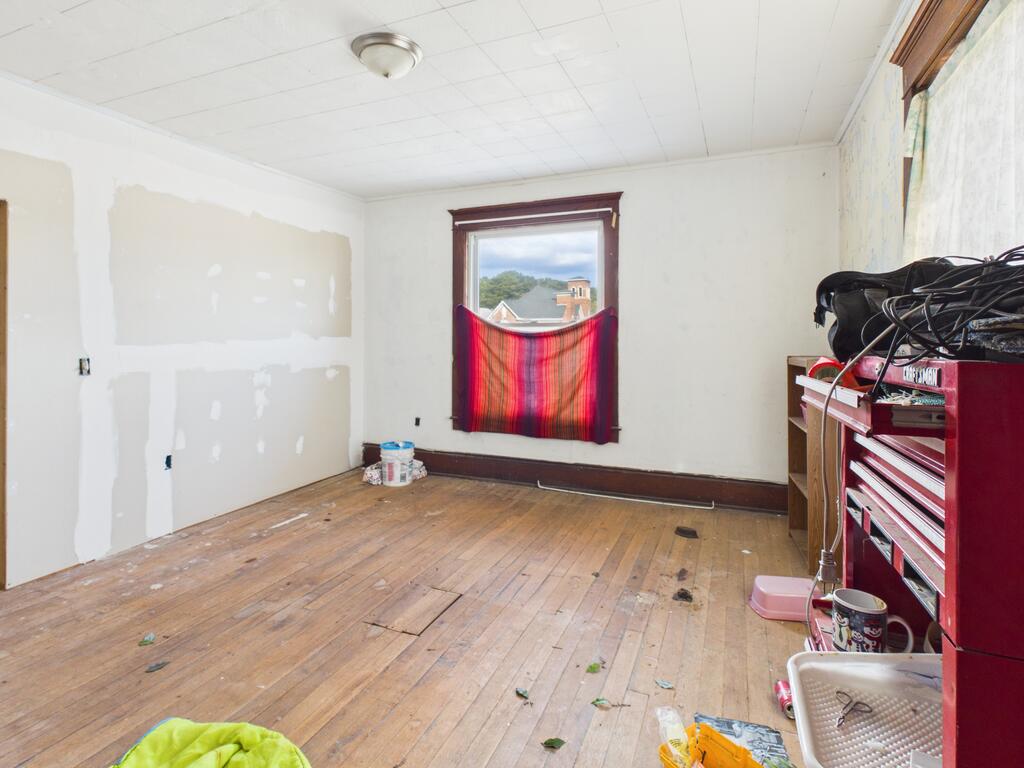 Room with worn hardwood floors, unfinished drywall, and a window covered by a red striped cloth.