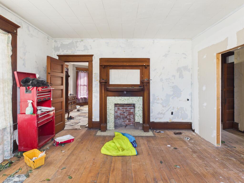 Vacant room with a wood-trimmed fireplace, peeling walls, and worn hardwood floors.