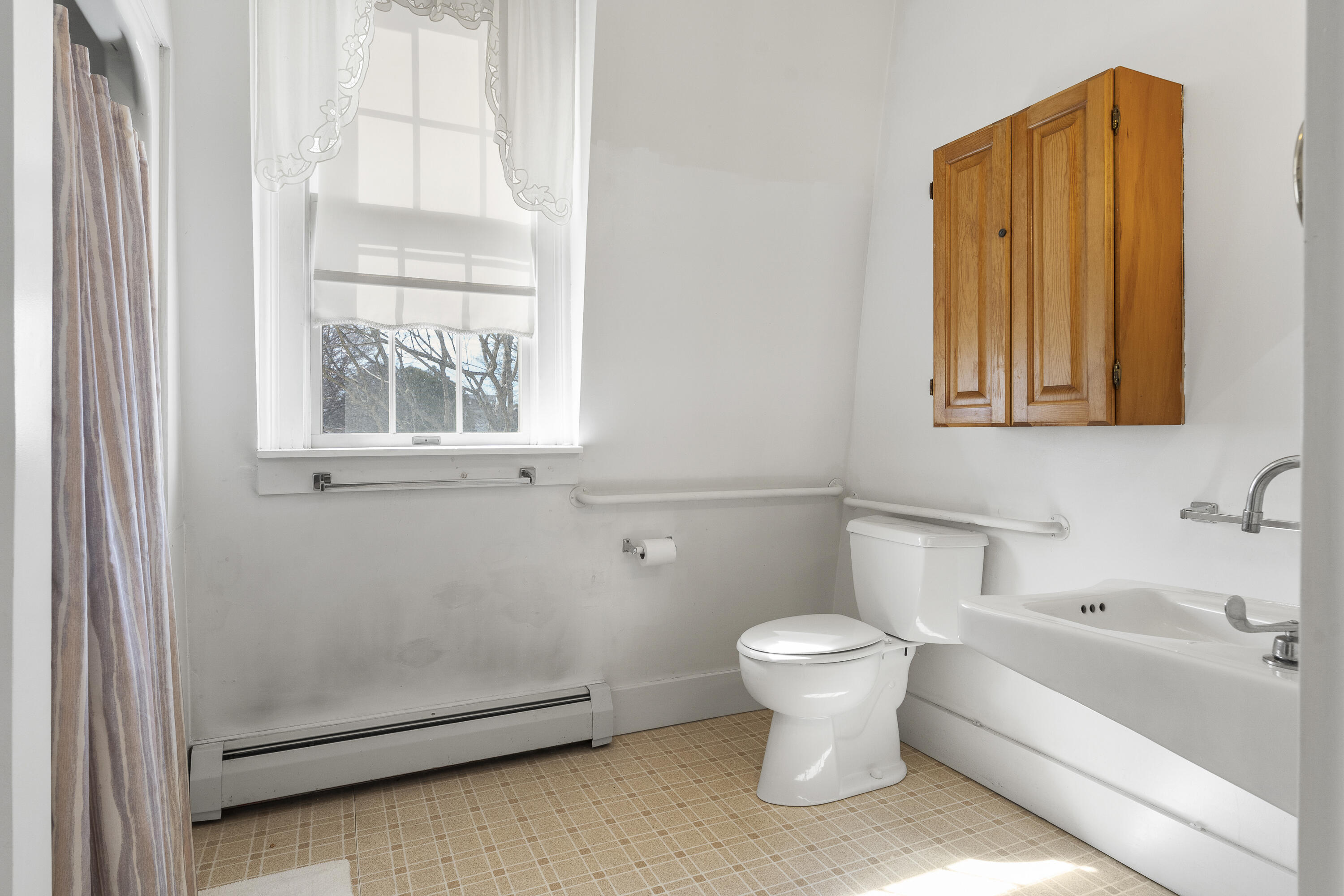 Bathroom with a bathtub, toilet, wooden cabinet, and grab bars beside a sunny window.