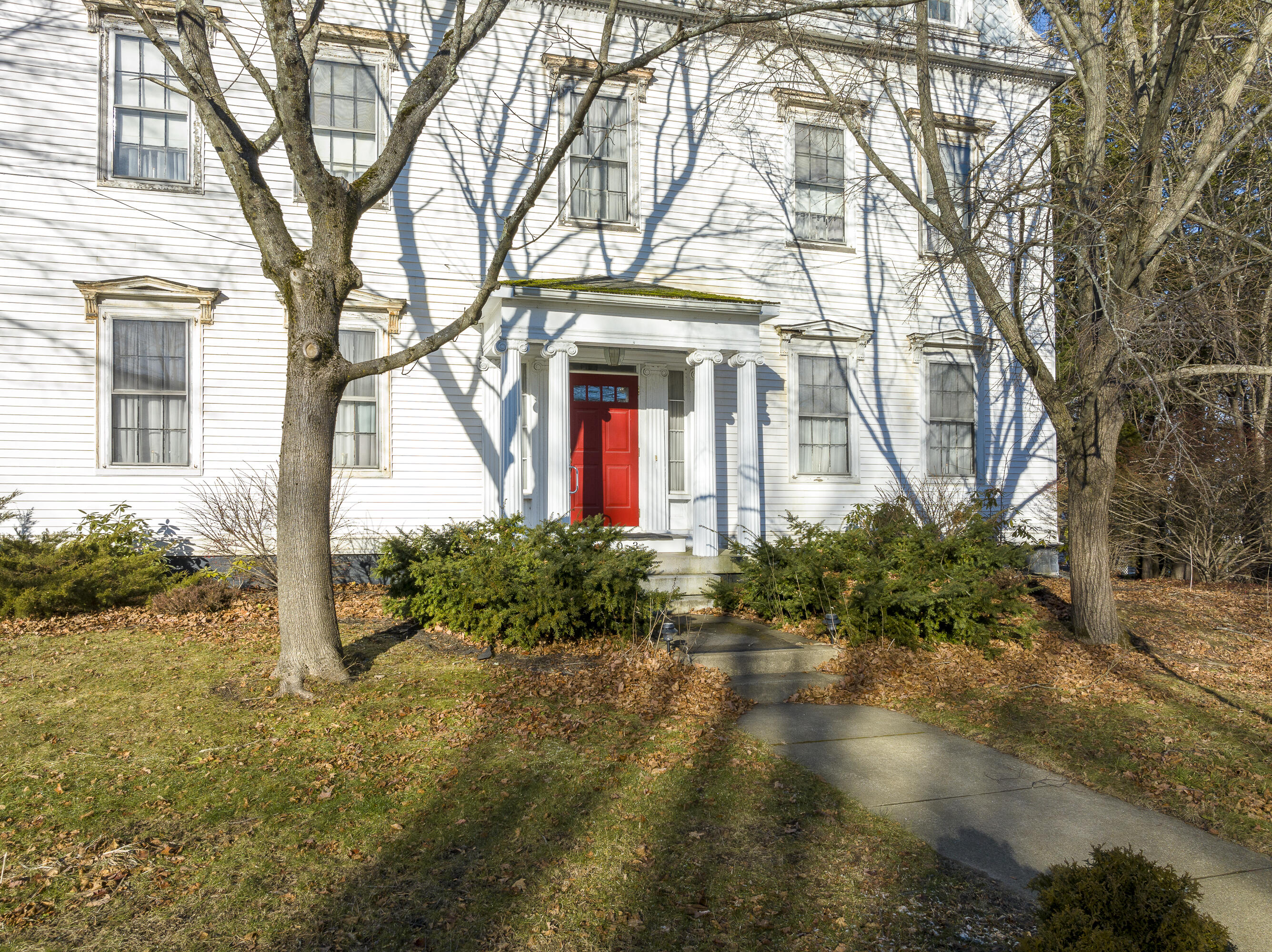 White clapboard house with a columned entrance and bright red door, framed by leafless trees.