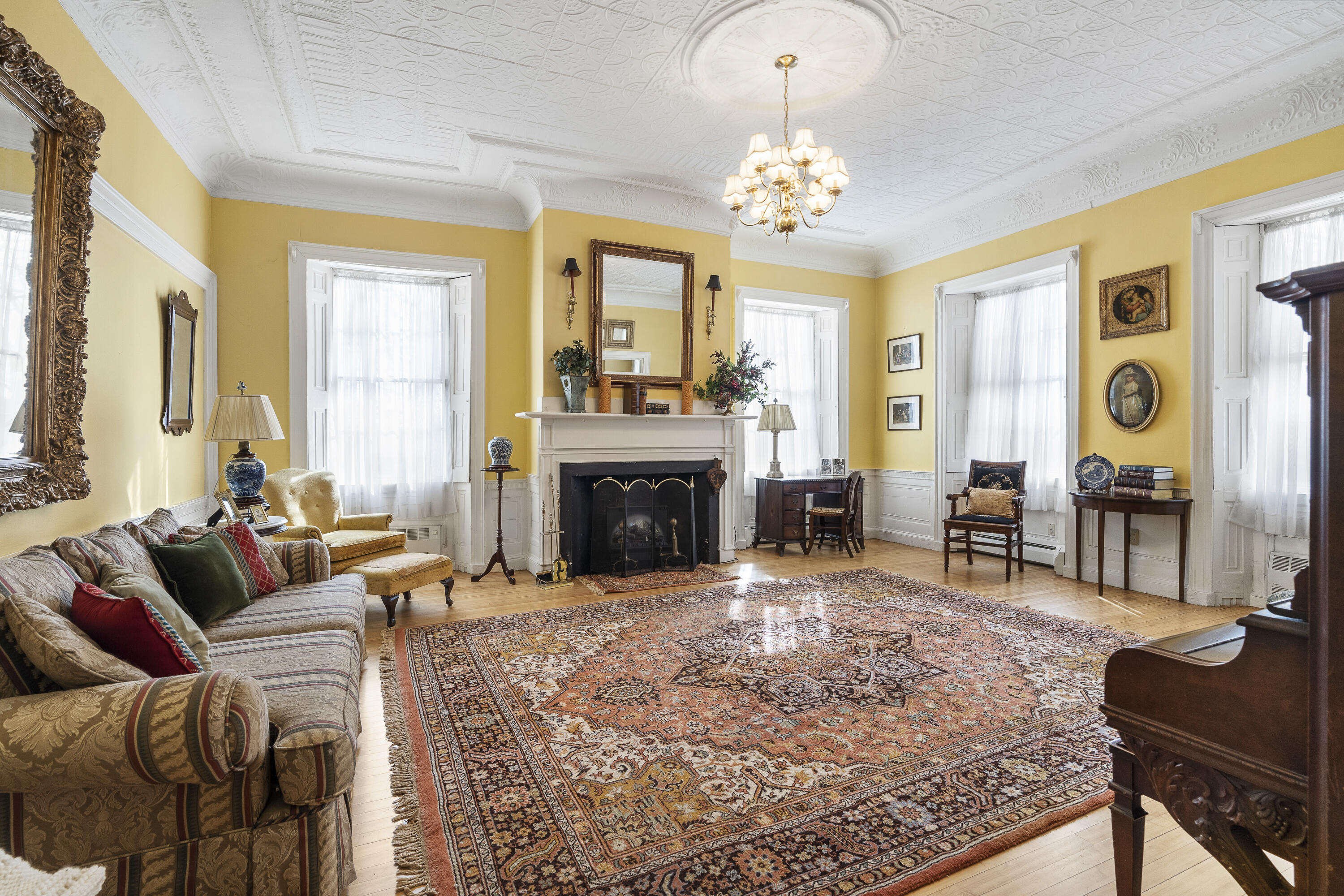 Living room with a white fireplace, ornate ceiling, tall windows, and a large patterned rug.
