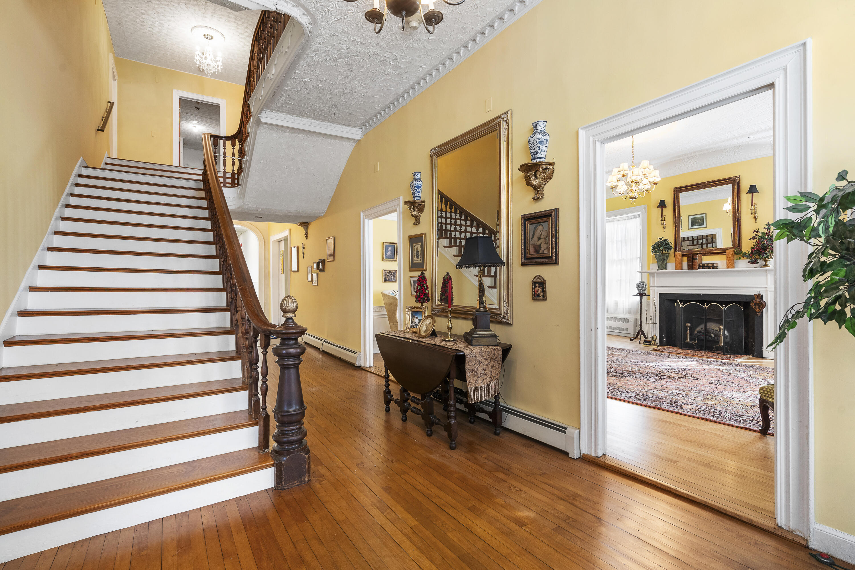 Historic foyer with a sweeping wood-trimmed staircase, hardwood floors, and a view into a fireplace room.