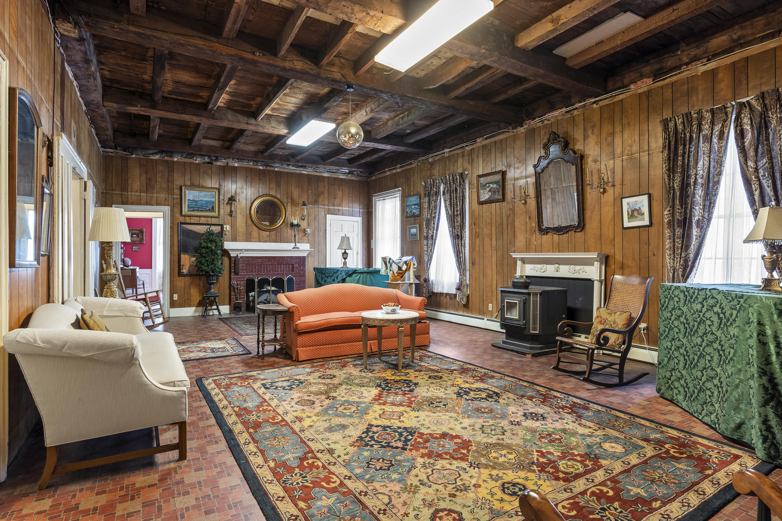 Wood-paneled living room with exposed beams, brick floors, two fireplaces, and a large patterned rug.