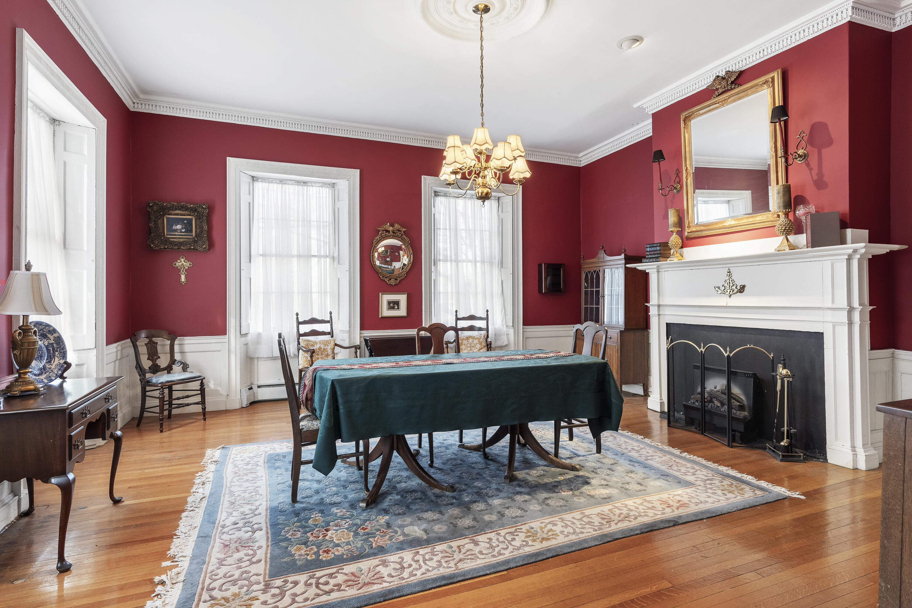 Dining room with red walls, an ornate white fireplace, chandelier, and dark-covered table.