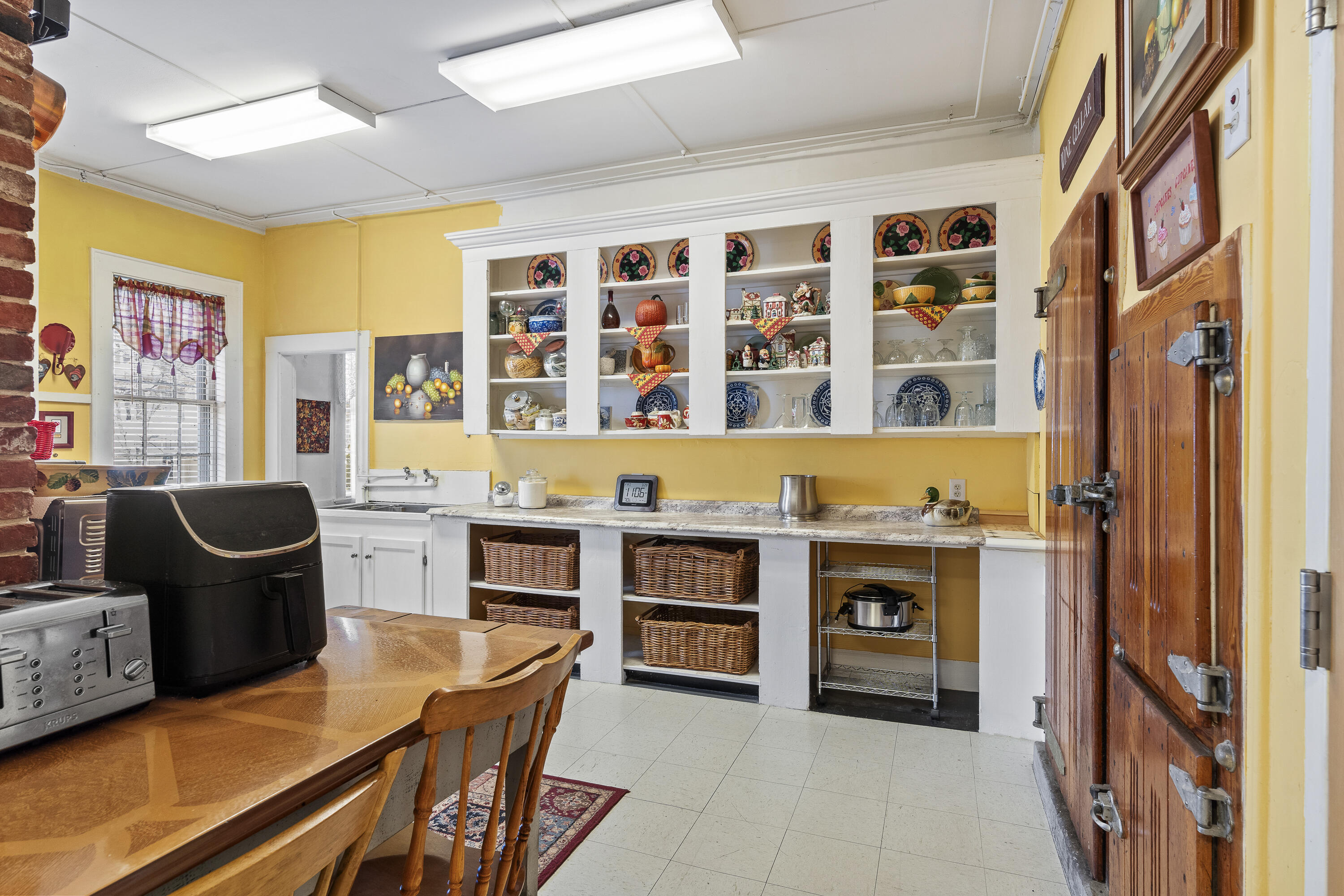 Kitchen with yellow walls, plate-filled white shelves, a wooden dining table, and a rustic wooden door.