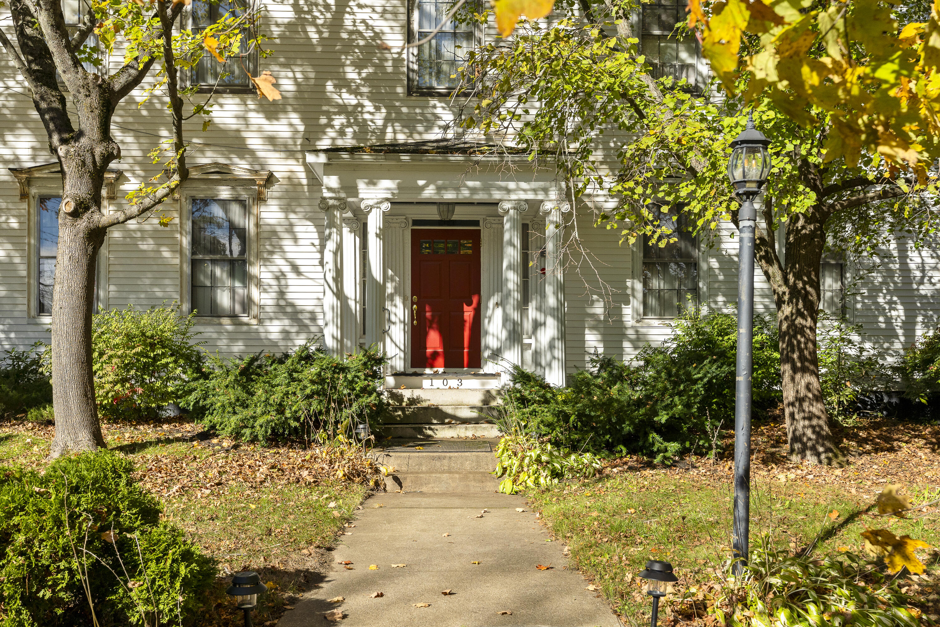 White clapboard house with a red front door, decorative columns, and a leaf-strewn garden path.