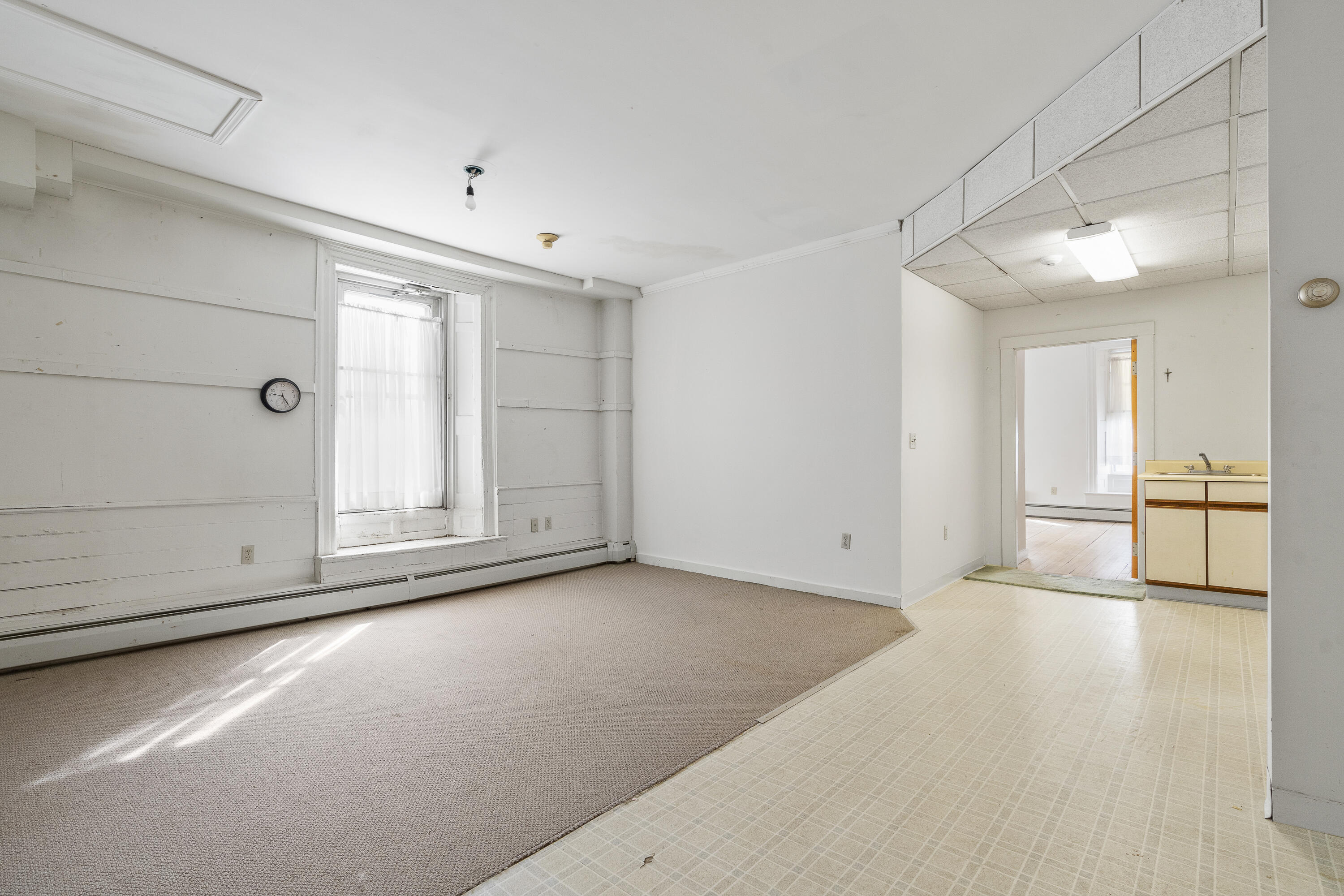 Empty room with a large curtained window and adjoining kitchenette.