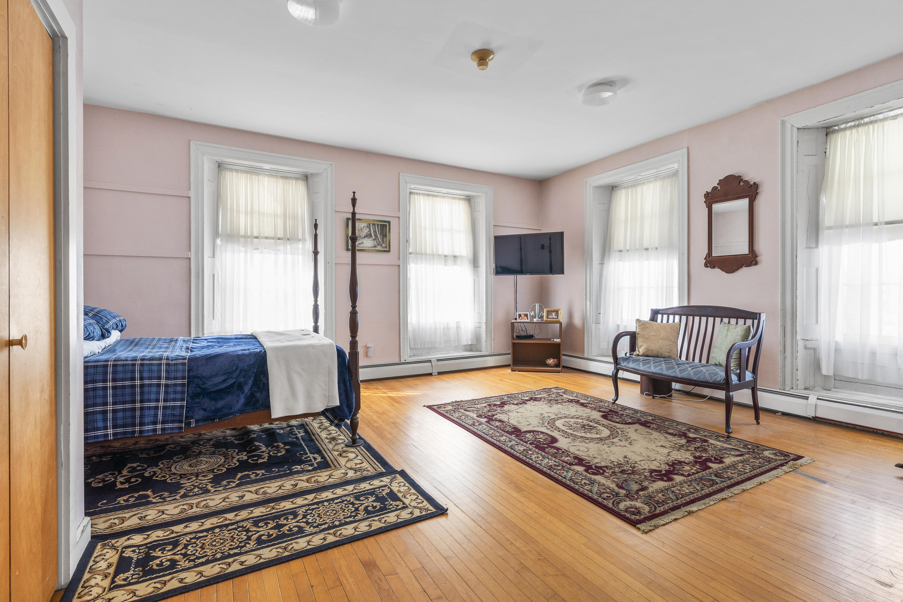 Bedroom with pink walls, a four-poster bed, tall curtained windows, and hardwood floors.