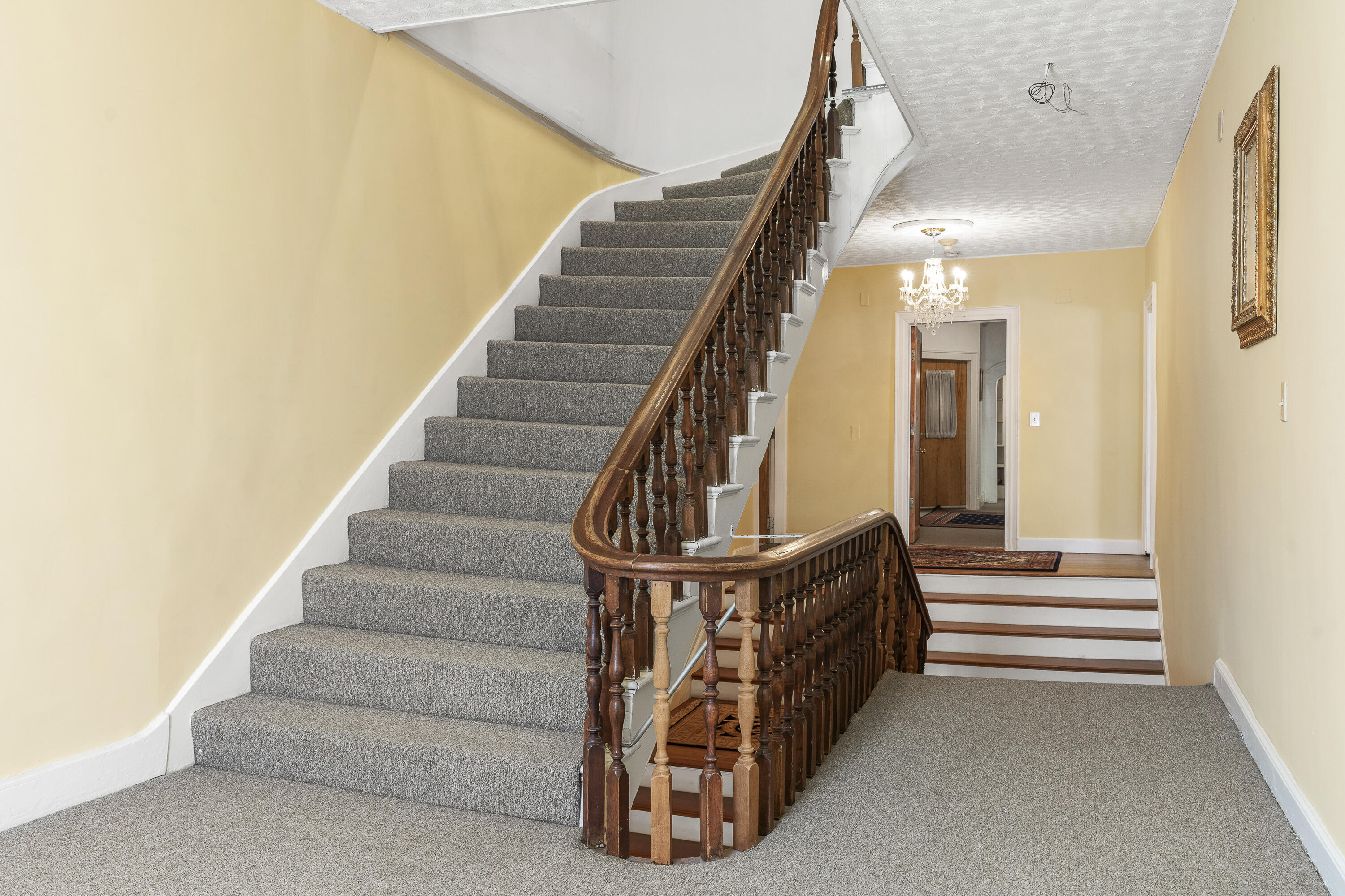 Stair hall with gray-carpeted stairs, an ornate wood banister, and a chandelier.