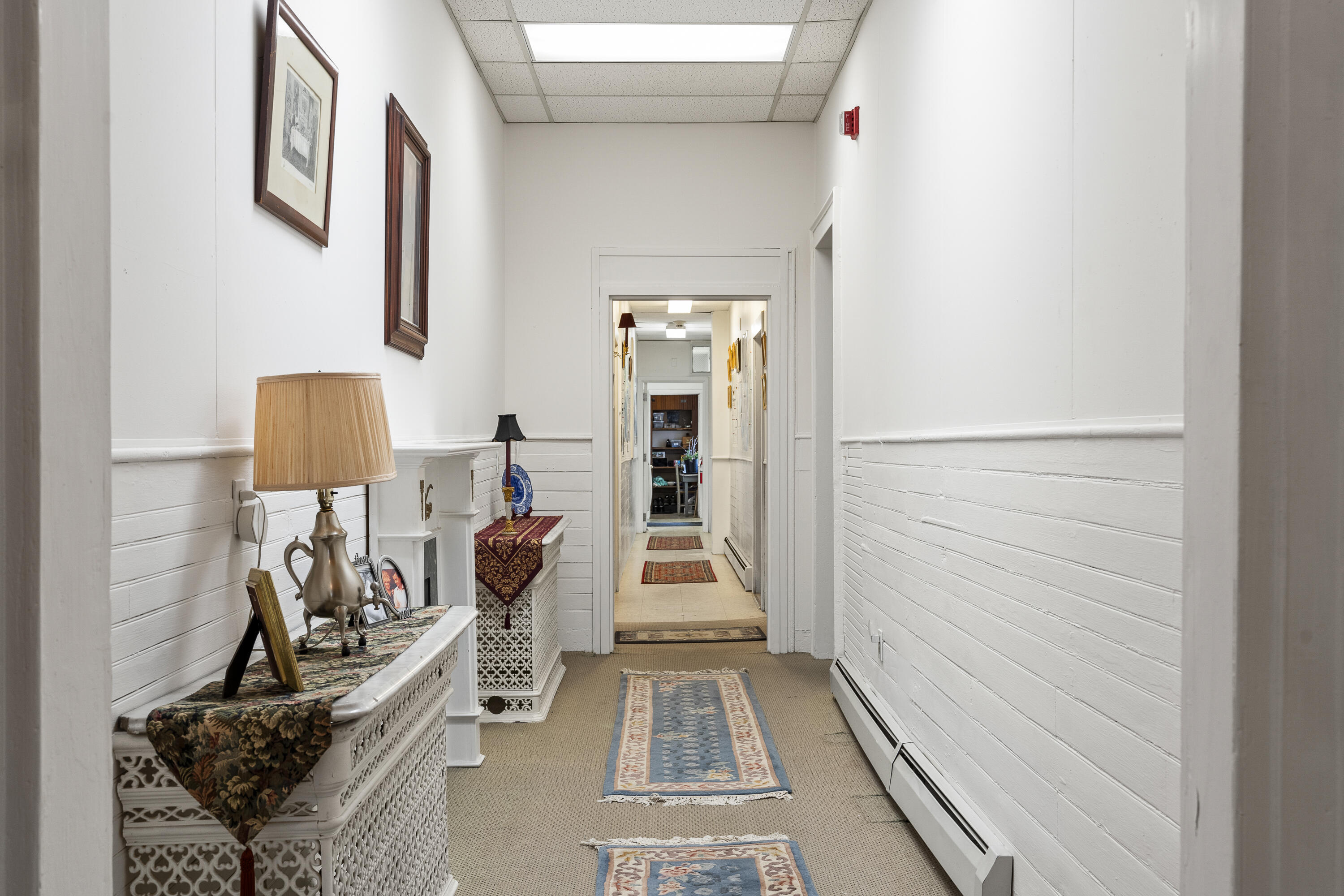 Furnished hallway with white paneling, patterned rugs, framed art, and an ornate radiator cover.