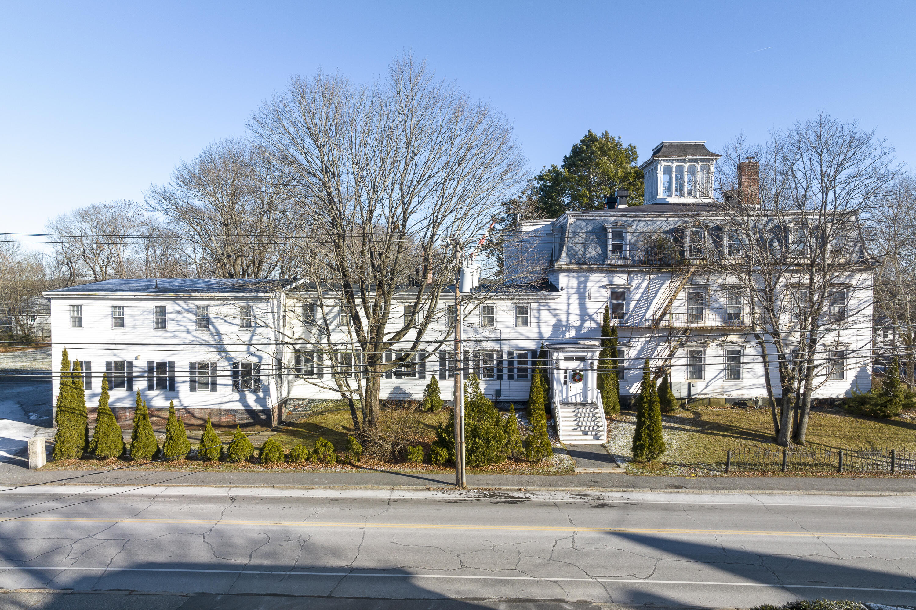 Large white historic house with a cupola, front steps, evergreen shrubs, and leafless trees.