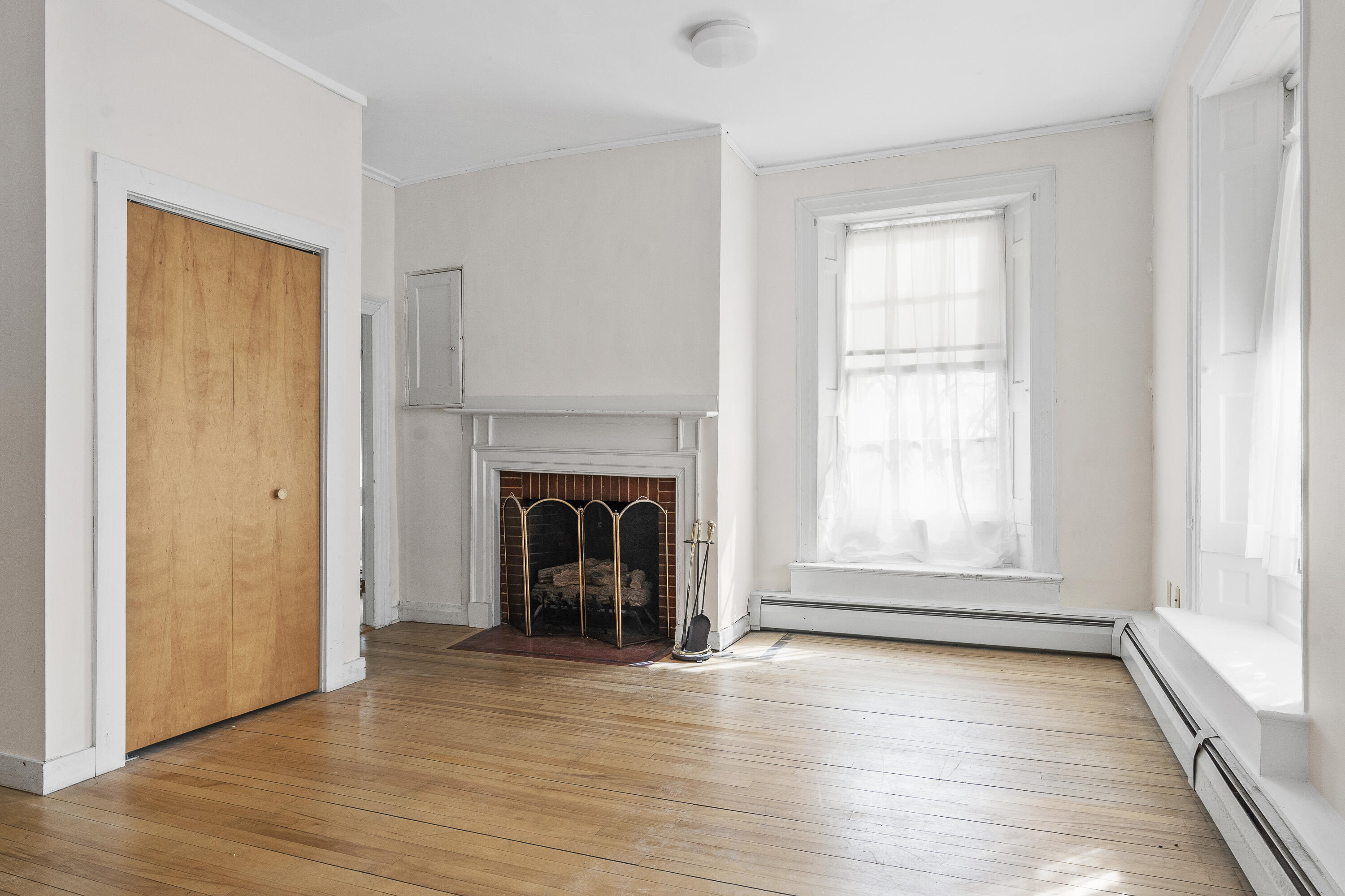 Empty room with hardwood floors, a brick fireplace, and tall windows with sheer curtains.