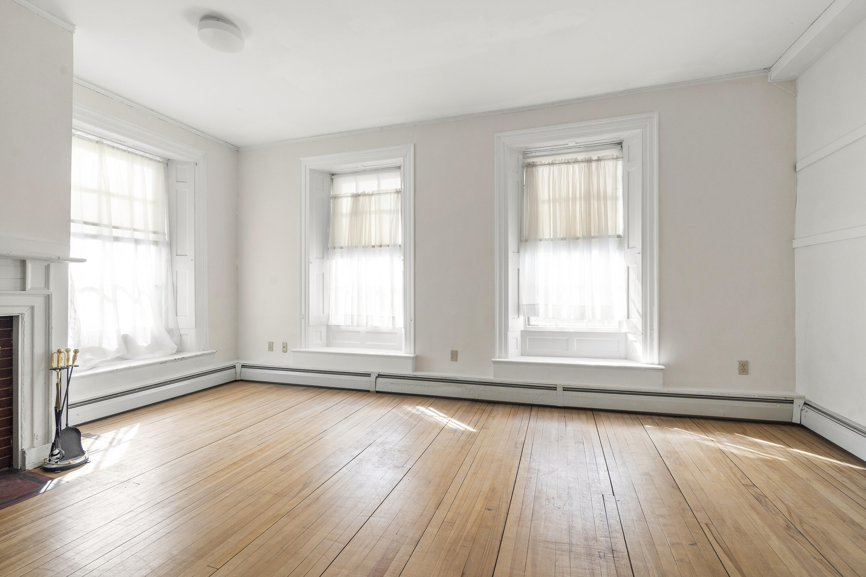 Bright vacant room with hardwood floors, three tall curtained windows, and a fireplace.
