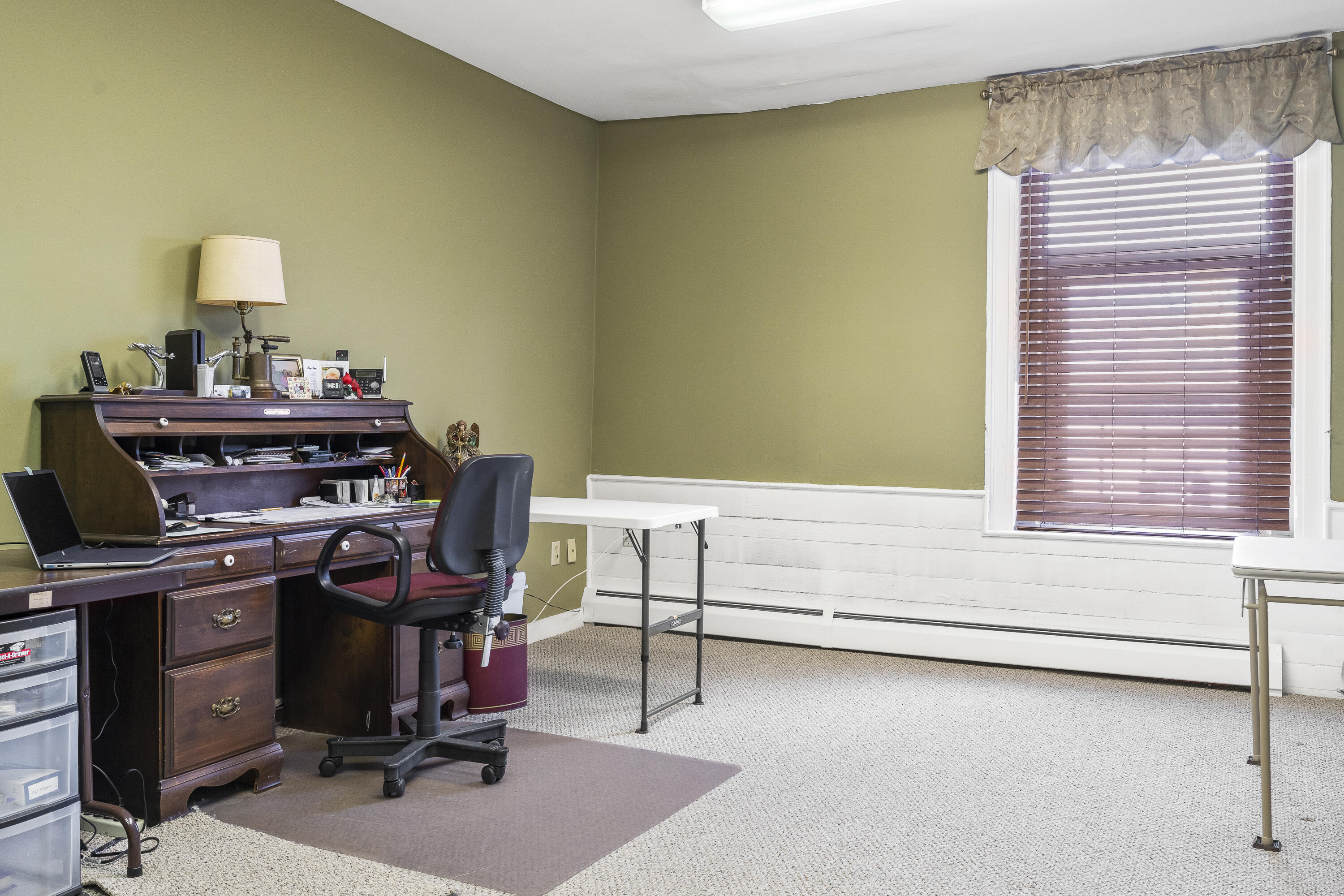 Furnished office with a dark wood roll-top desk, olive-green walls, and a large window with blinds.