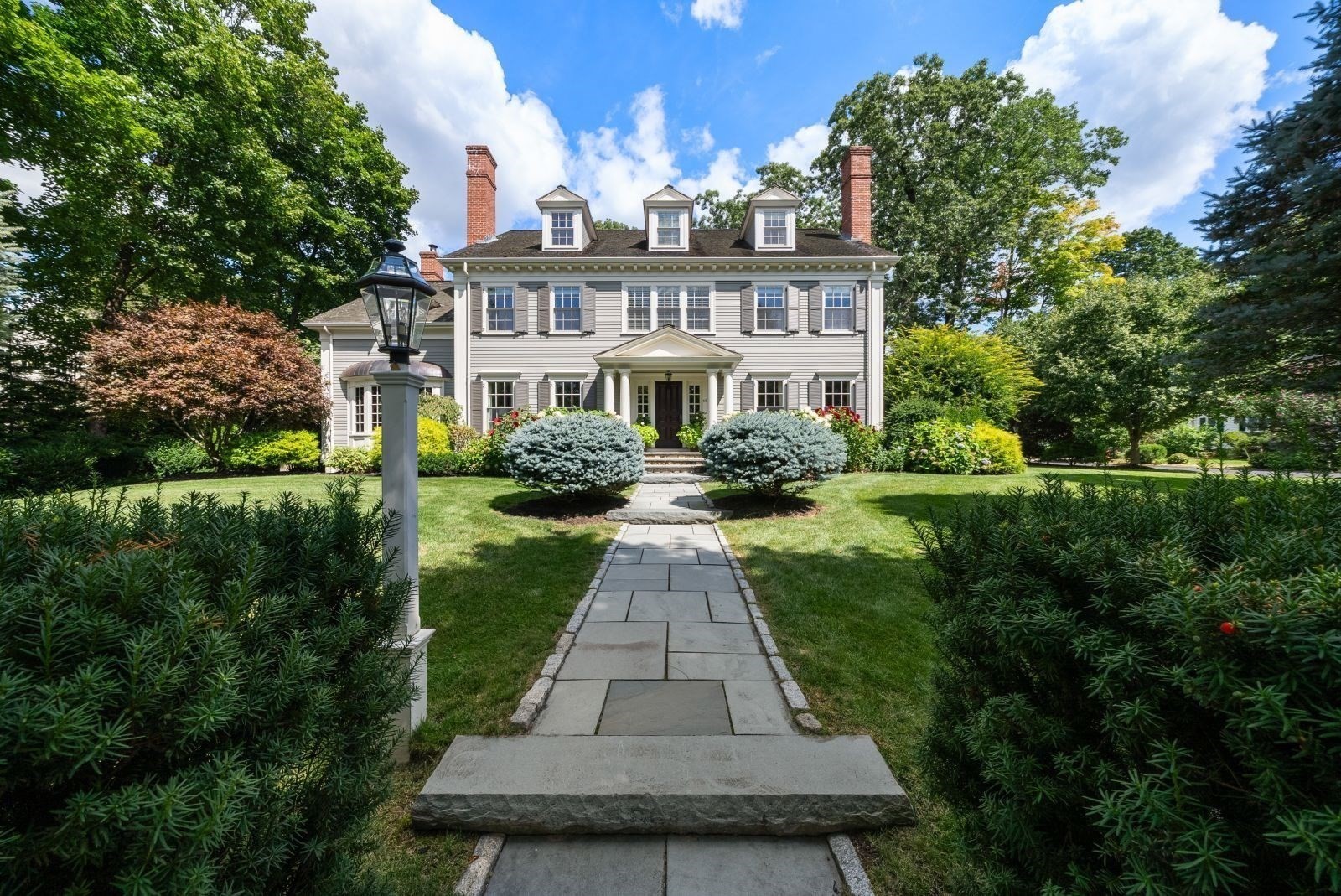 White Colonial home with three dormers, brick chimneys, and a stone walkway through landscaped gardens.
