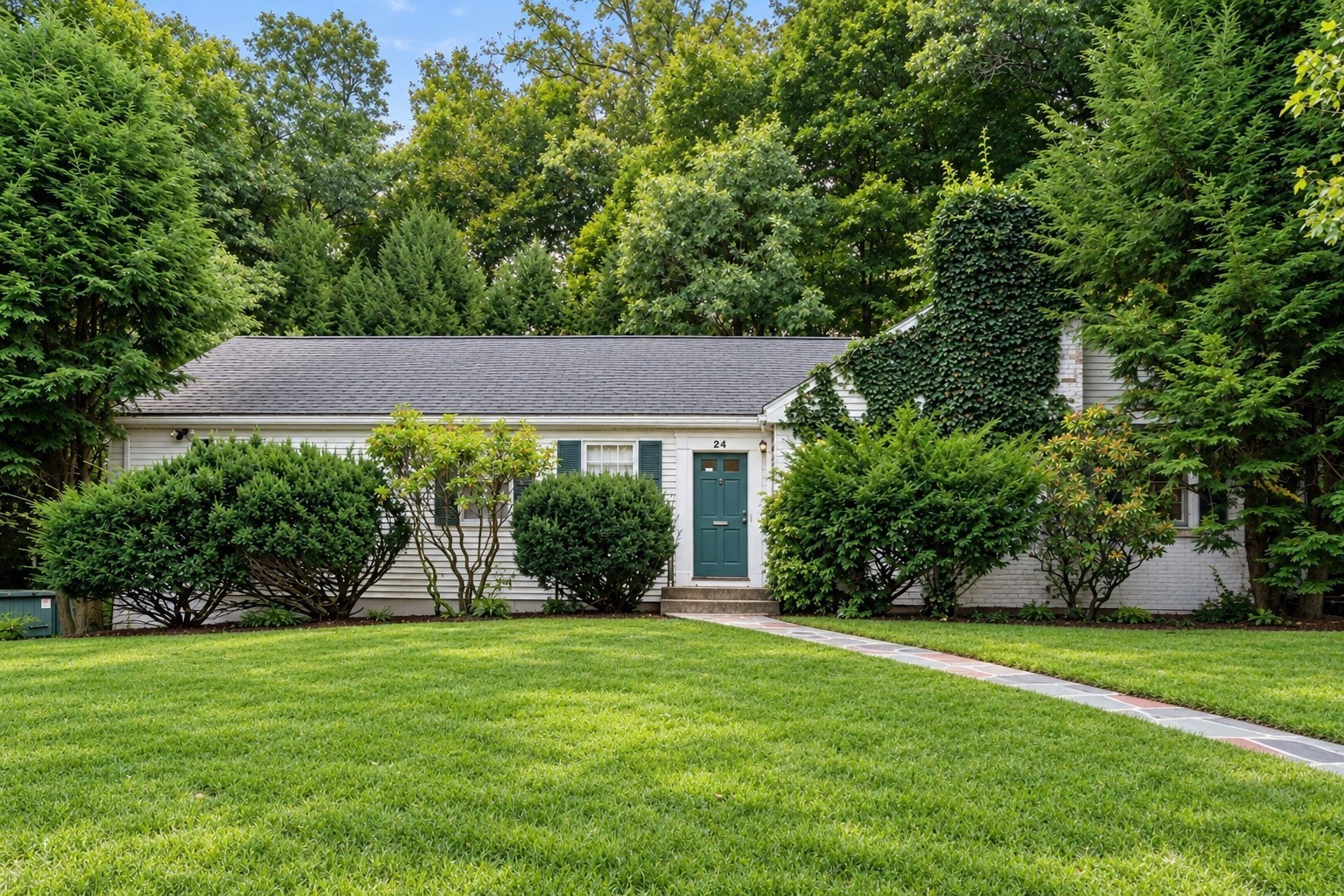 Single-story white house with a teal front door, shaded by mature trees and surrounded by lush landscaping.