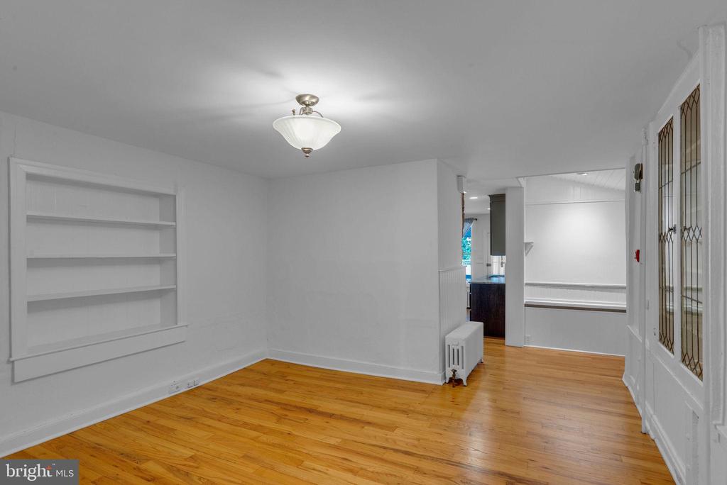 Vacant room with hardwood floors, built-in shelving, and a doorway toward the kitchen.