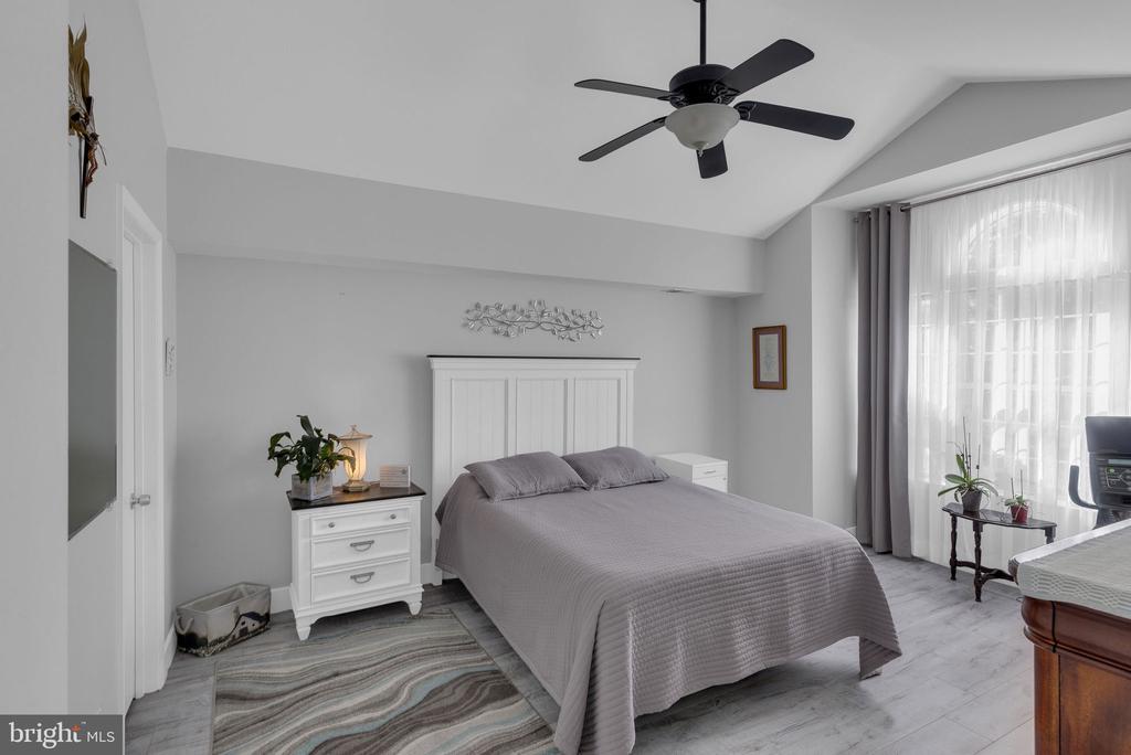 Bedroom with a gray bed, white paneled headboard, ceiling fan, and large curtained window.