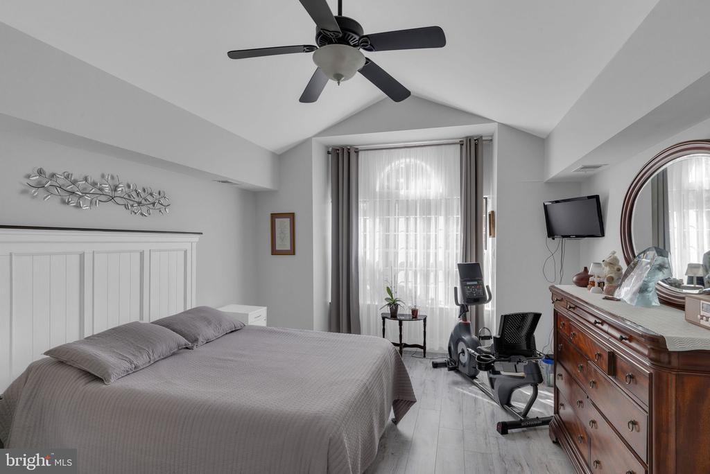 Bedroom with a gray bed, vaulted ceiling, exercise bike, and wooden dresser with round mirror.