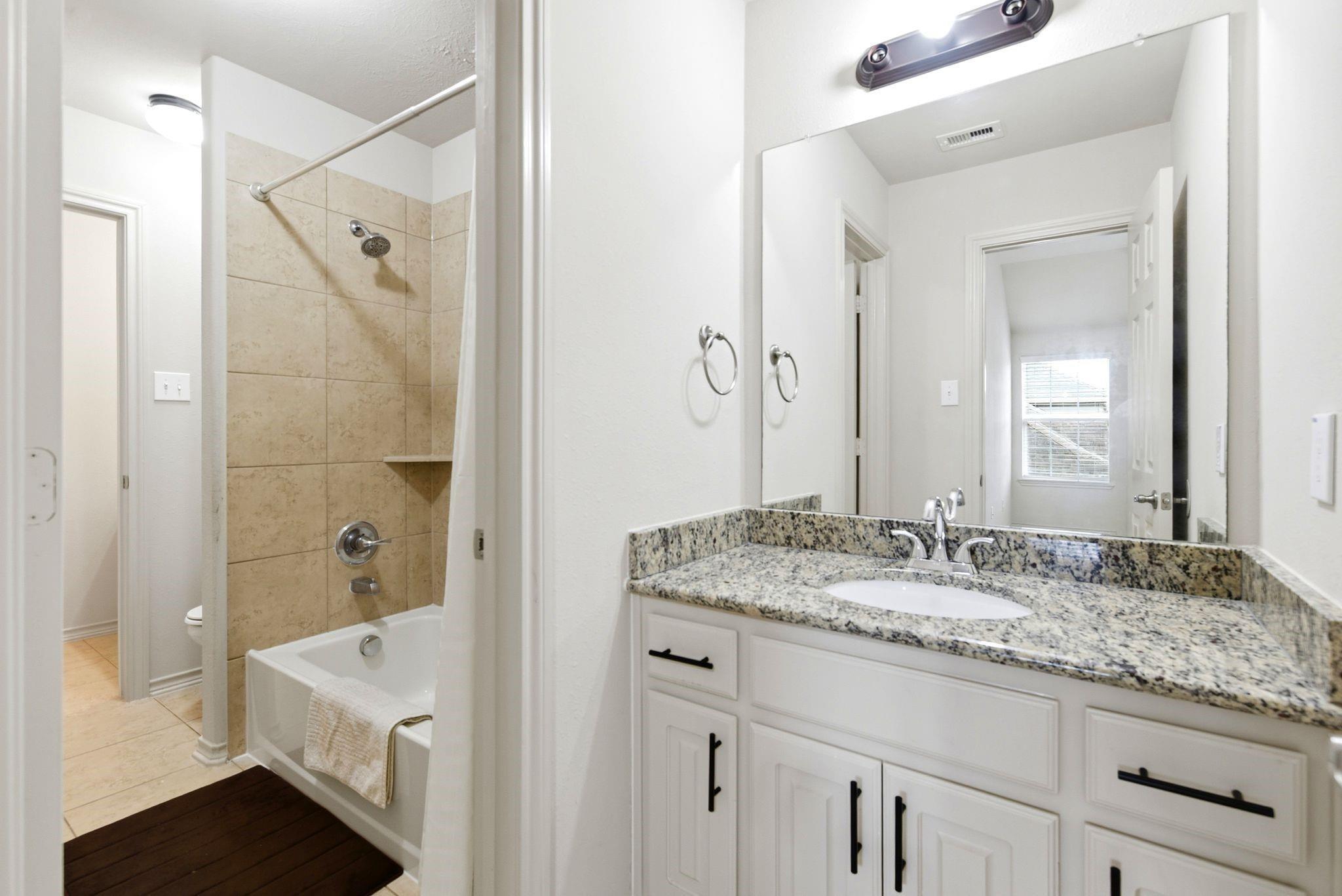 Bathroom with granite vanity and white cabinetry beside a beige tiled tub-shower.