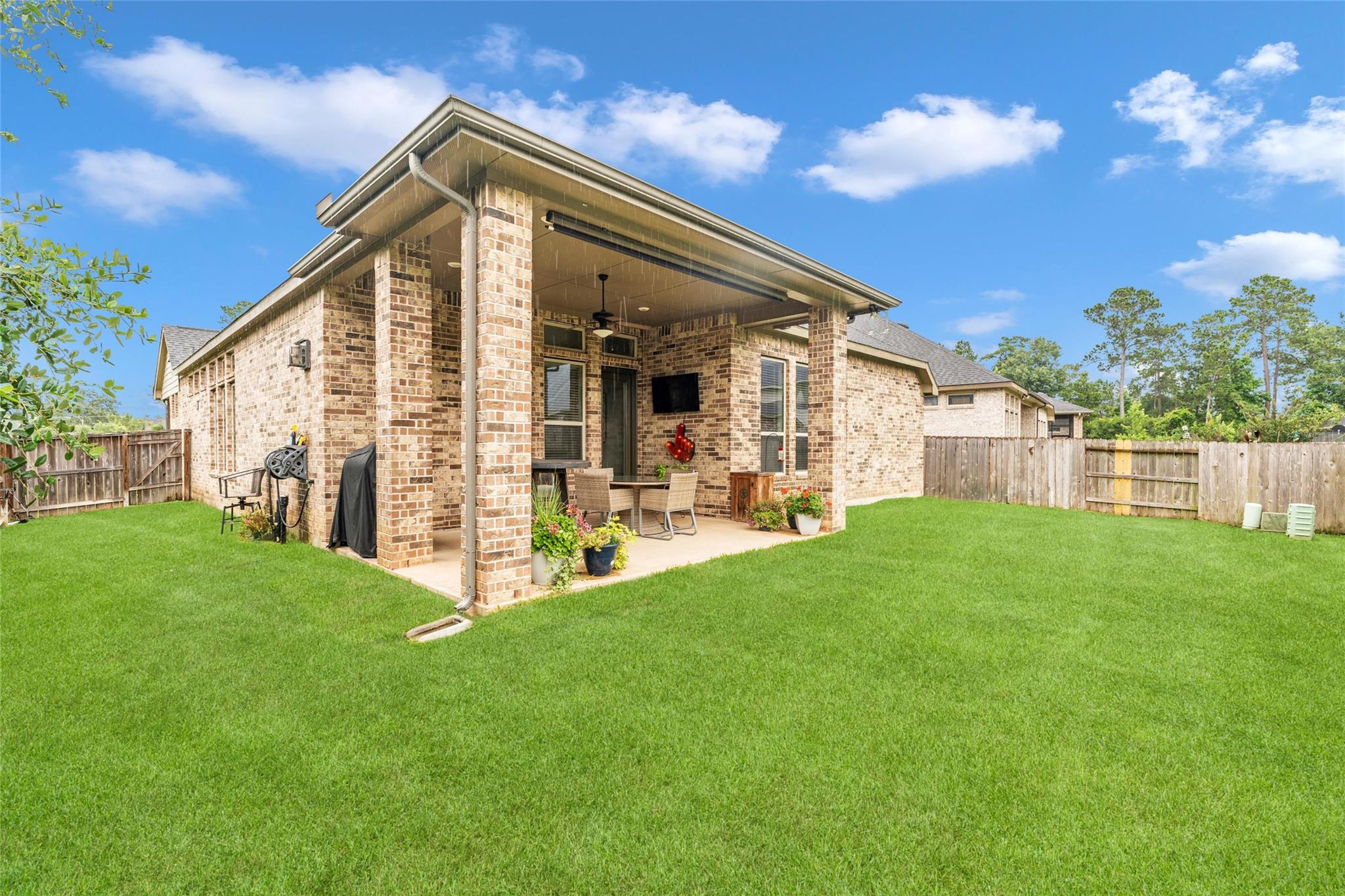 Brick home’s covered backyard patio with outdoor seating and potted flowers beside a broad green lawn.