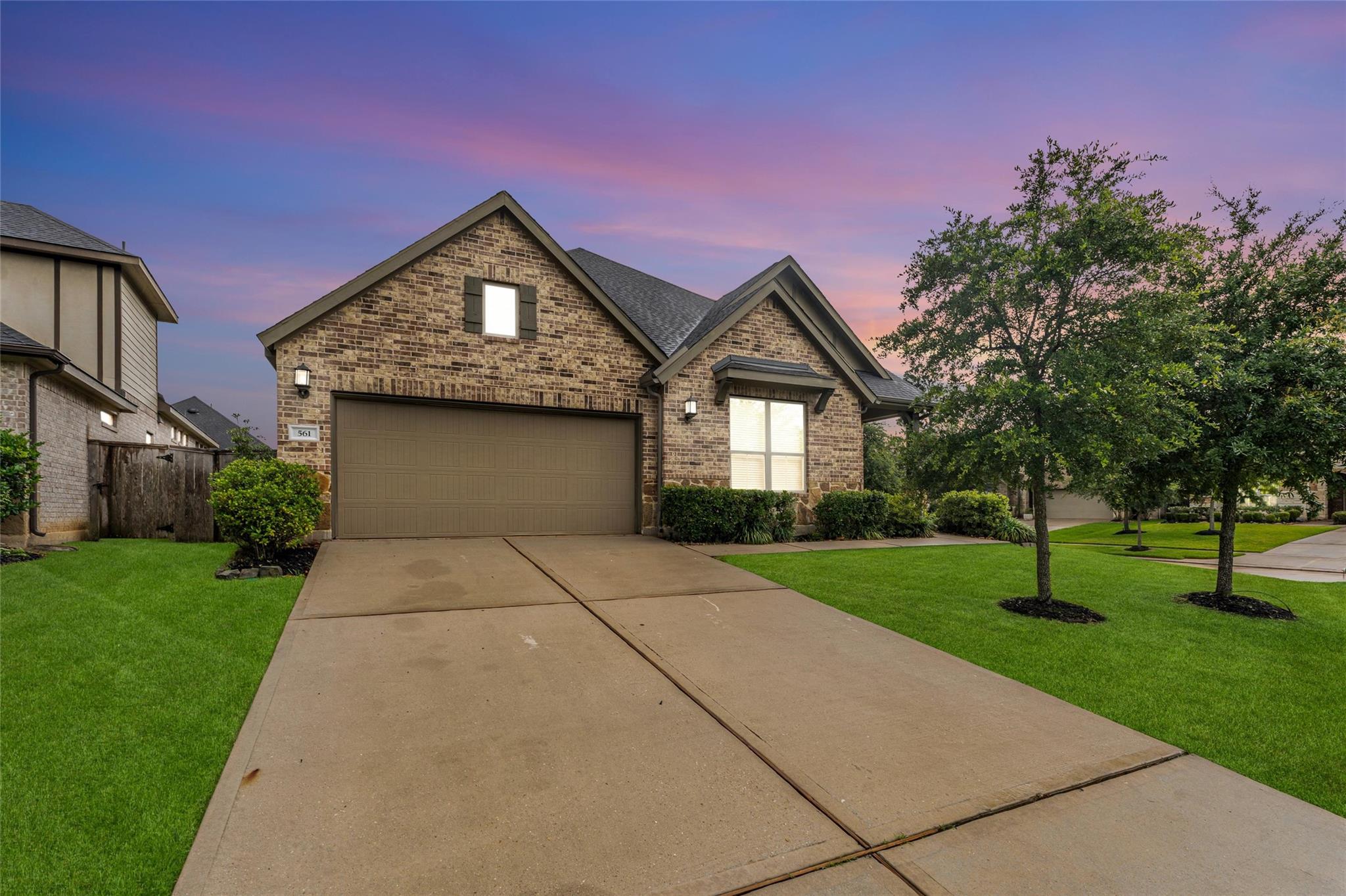 Brick home with a two-car garage, broad driveway, and landscaped front lawn at dusk.