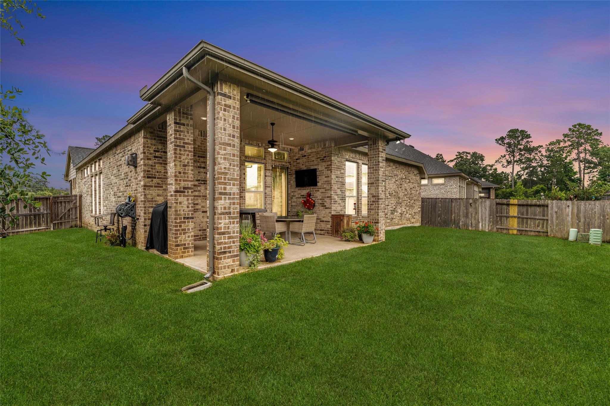 Brick home with a covered, furnished patio overlooking a fenced lawn at dusk.