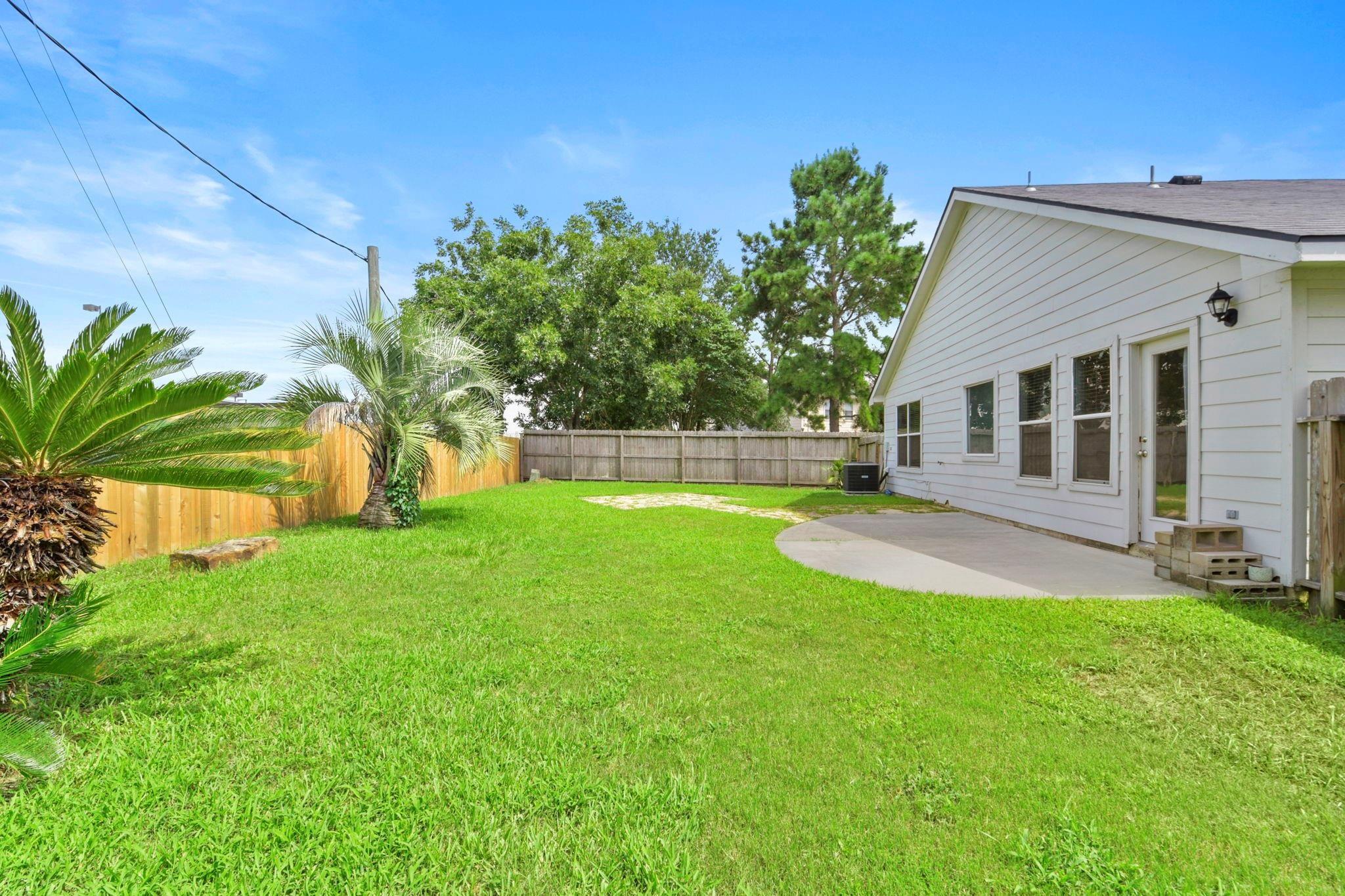Fenced backyard with a wide lawn, palm trees, and a curved patio beside the house.