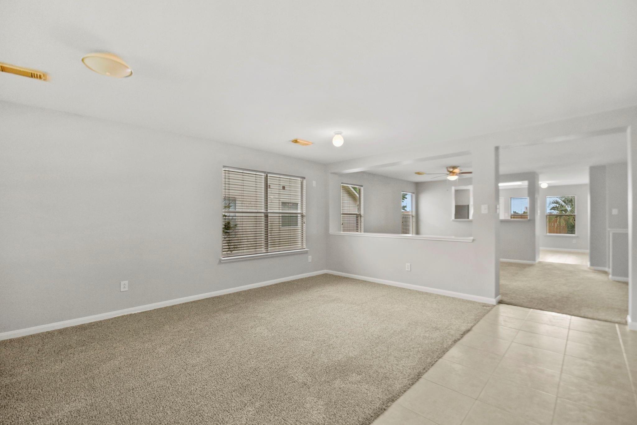 Empty living area with beige carpet, window blinds, and an open passage to adjoining rooms.