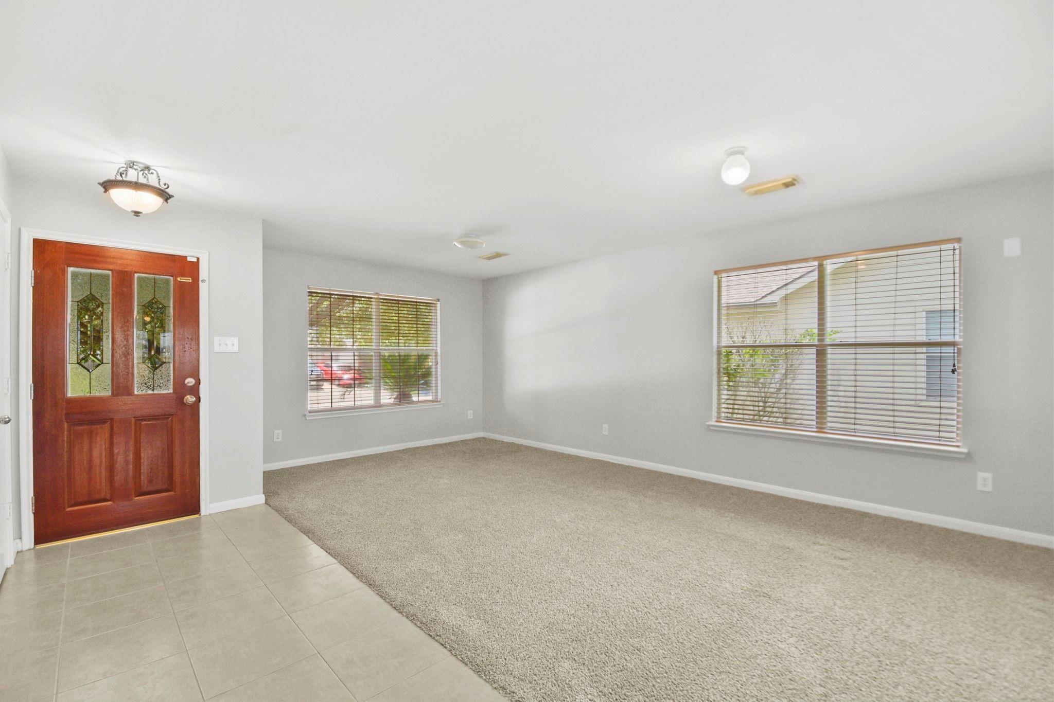 Vacant living room with beige carpet, two windows with blinds, and a dark wood front door.