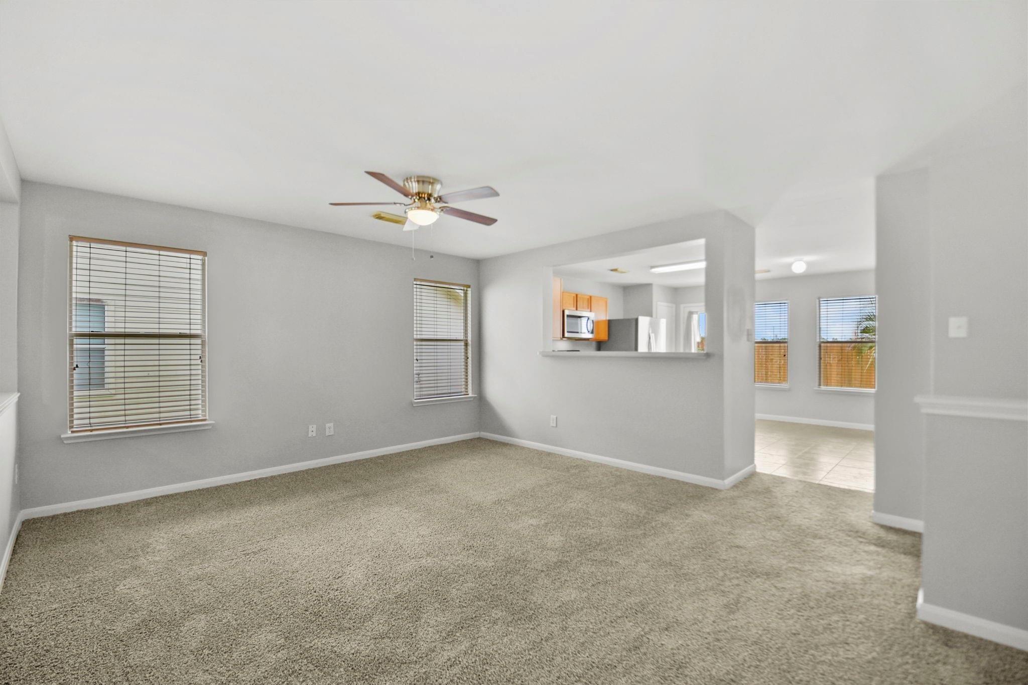 Empty carpeted living room with gray walls, ceiling fan, and an opening to the kitchen.