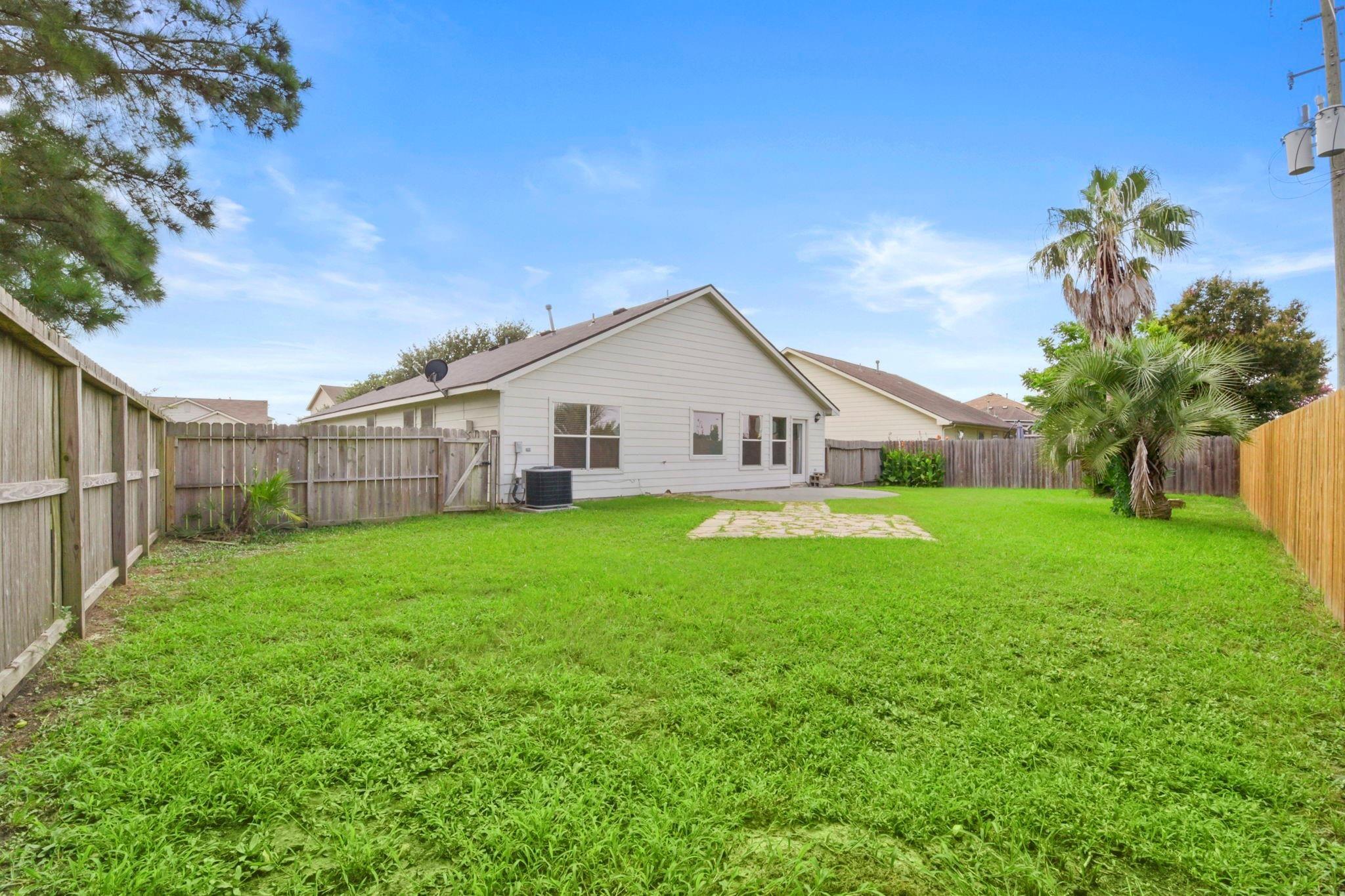 House viewed from a fenced backyard with a broad lawn, small patio, and palm trees.