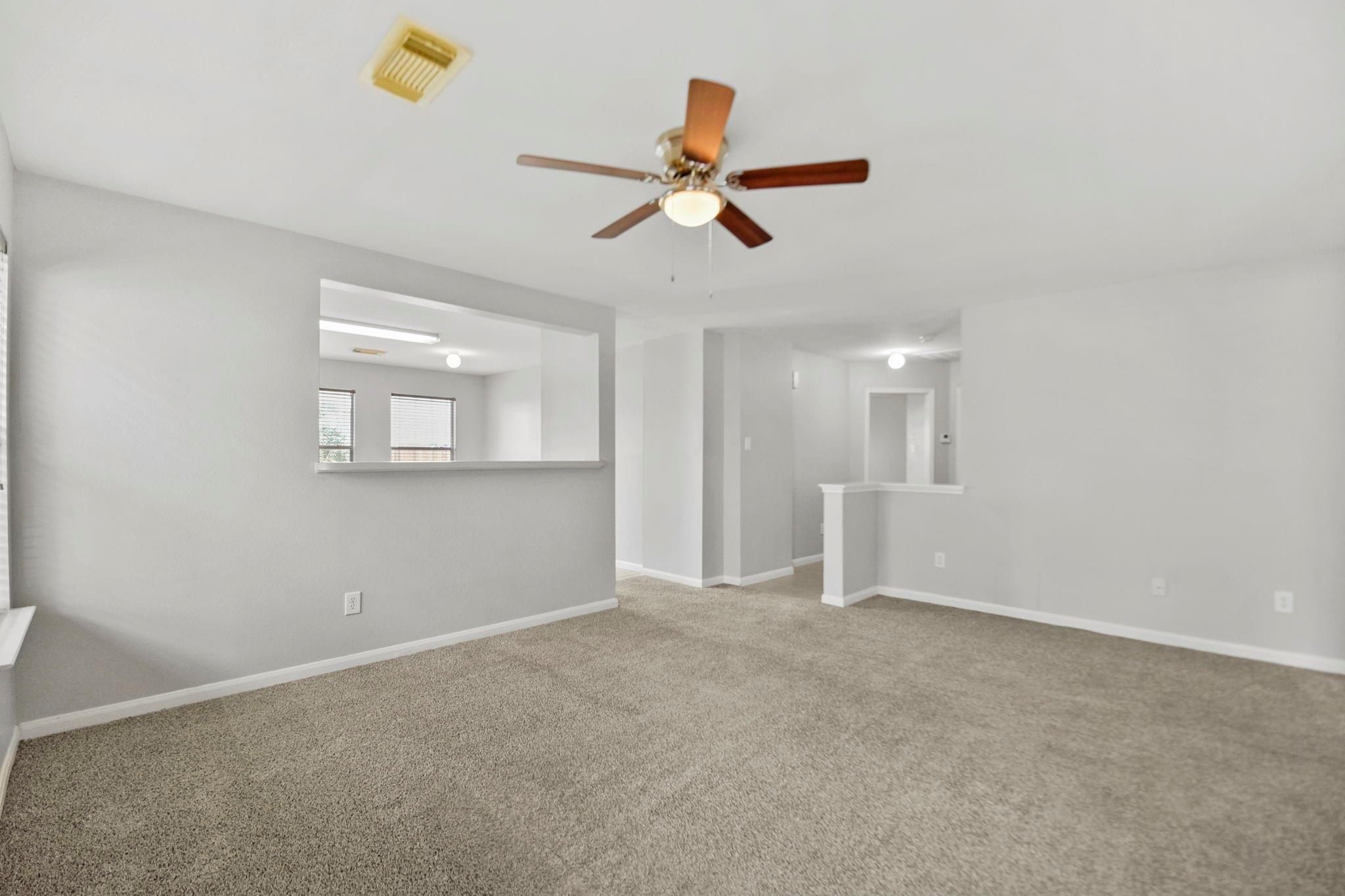 Empty carpeted living room with a ceiling fan and pass-through opening to an adjacent room.