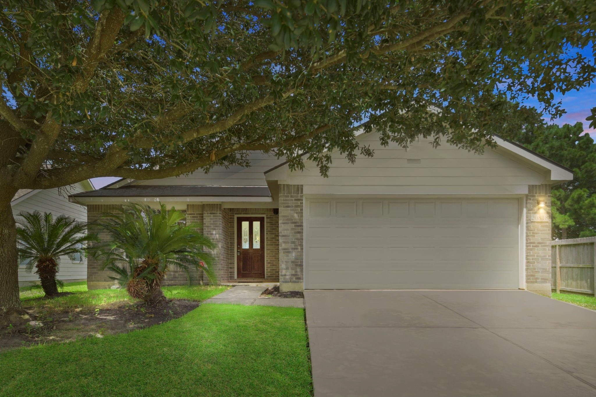 Brick home with a double garage, shaded by a large tree and bordered by palm landscaping.
