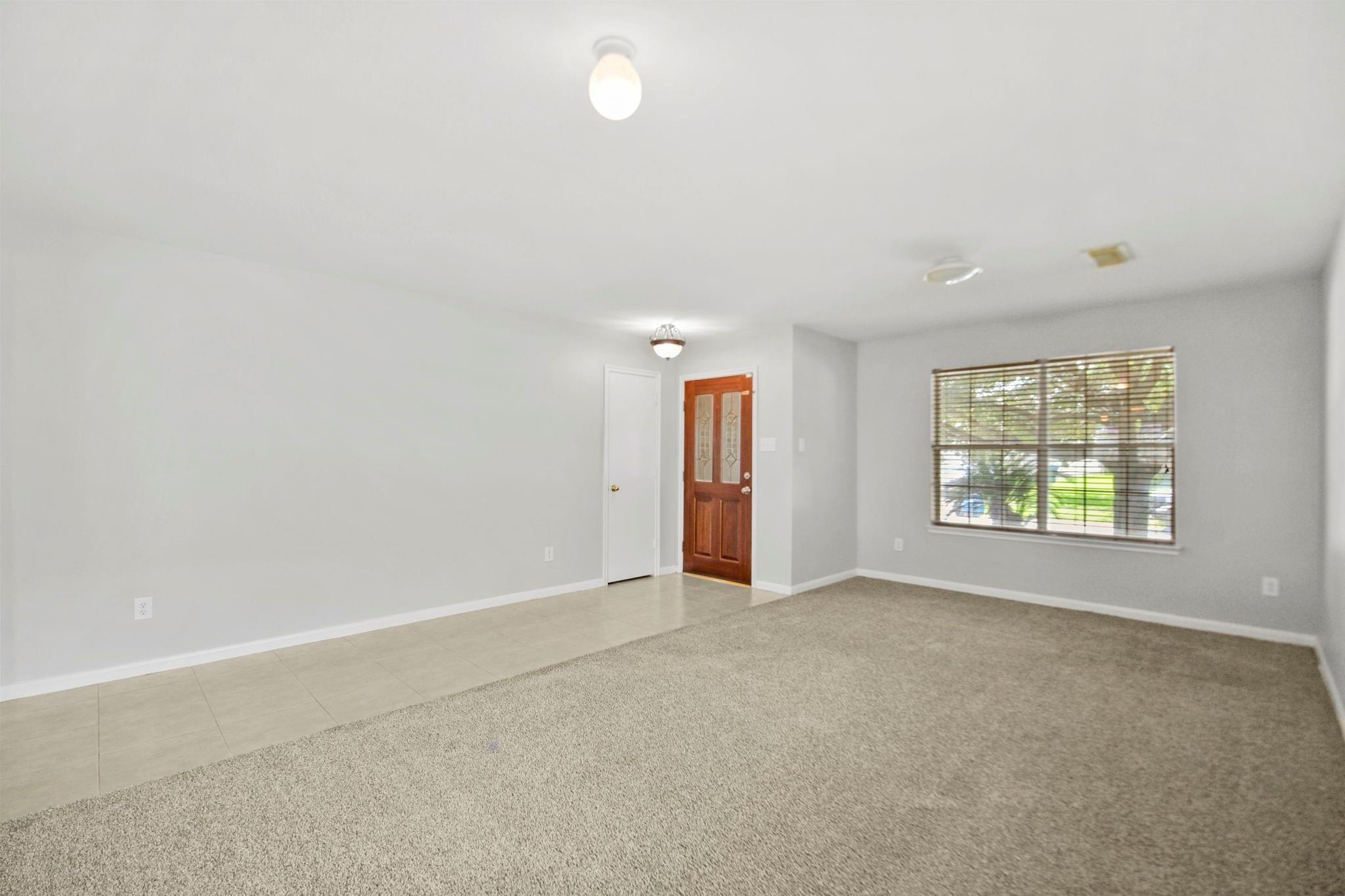 Empty living room with beige carpet, tiled entry, wood front door, and large window with blinds.