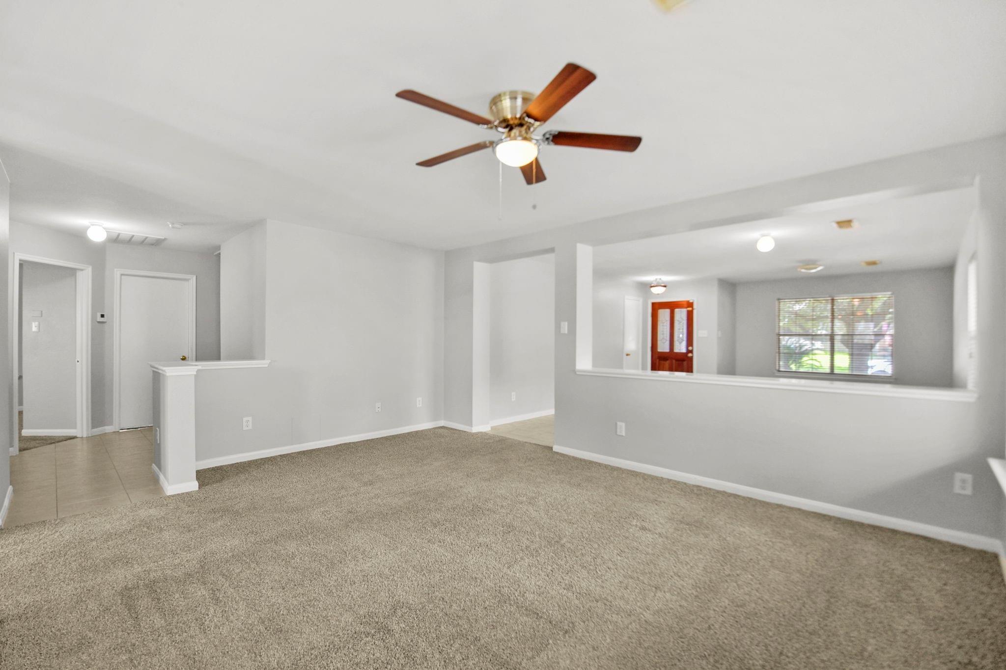 Empty living room with beige carpet, a wood-blade ceiling fan, and an open view toward the entry.