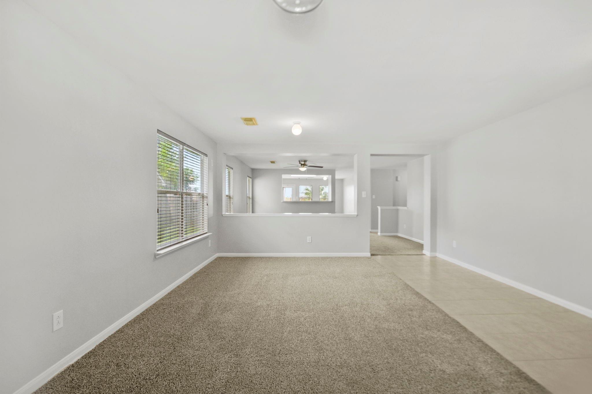 Empty carpeted living room with window blinds, a wide pass-through, and an adjoining tiled walkway.