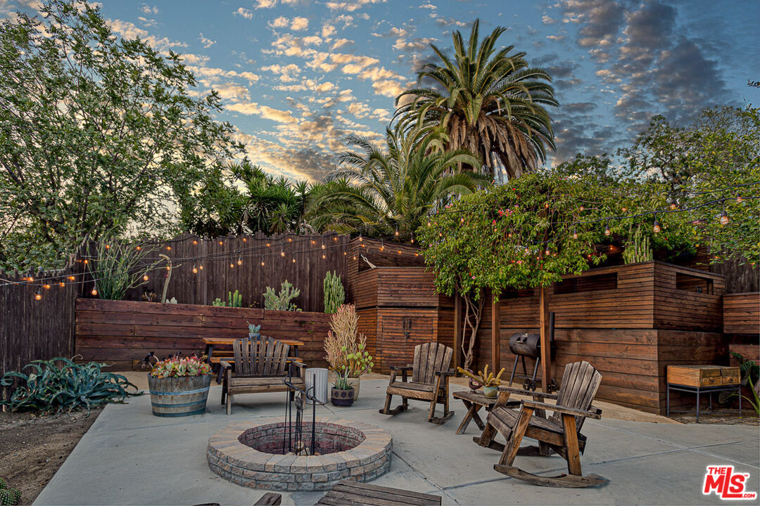 Backyard patio with a circular stone fire pit, wooden chairs, and string lights.