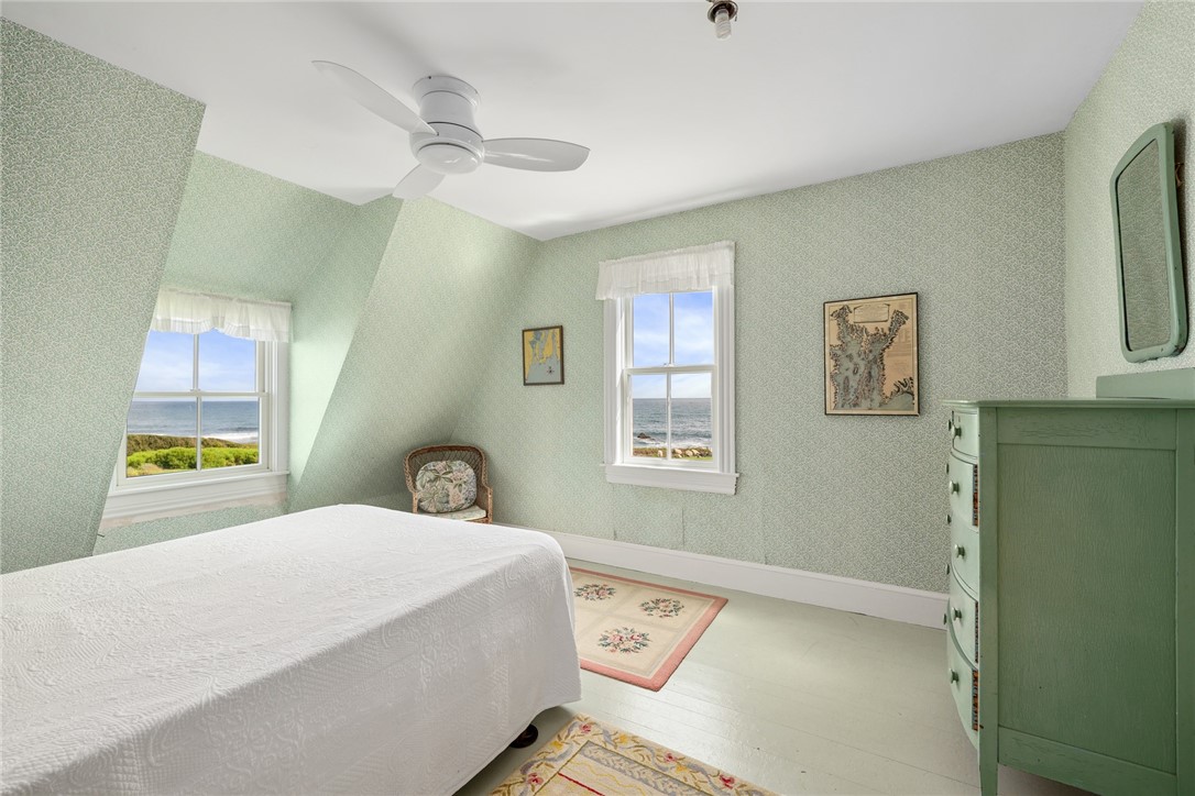 Bedroom with pale green walls, a white bed, vintage dresser, and ocean views through two windows.