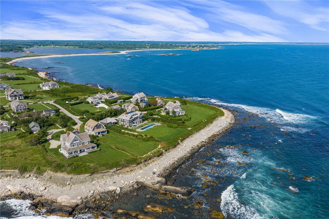 Coastal homes and green lawns along a rocky shore beside a broad blue bay.