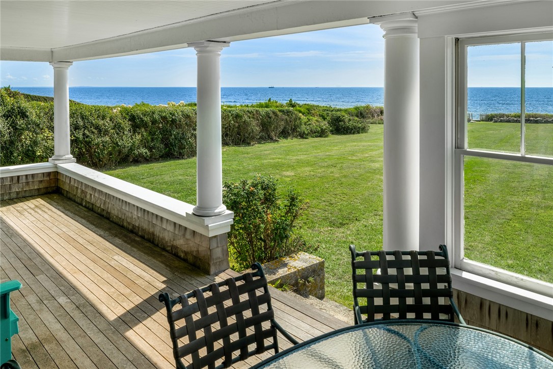 Covered porch with white columns and black chairs overlooking a lawn and the ocean.
