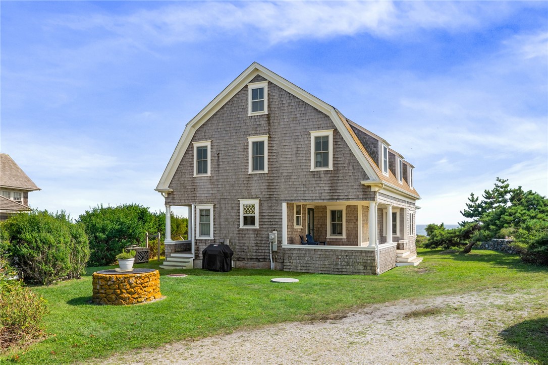 Gray shingle coastal house with a covered porch, stone well, and water visible beyond the lawn.