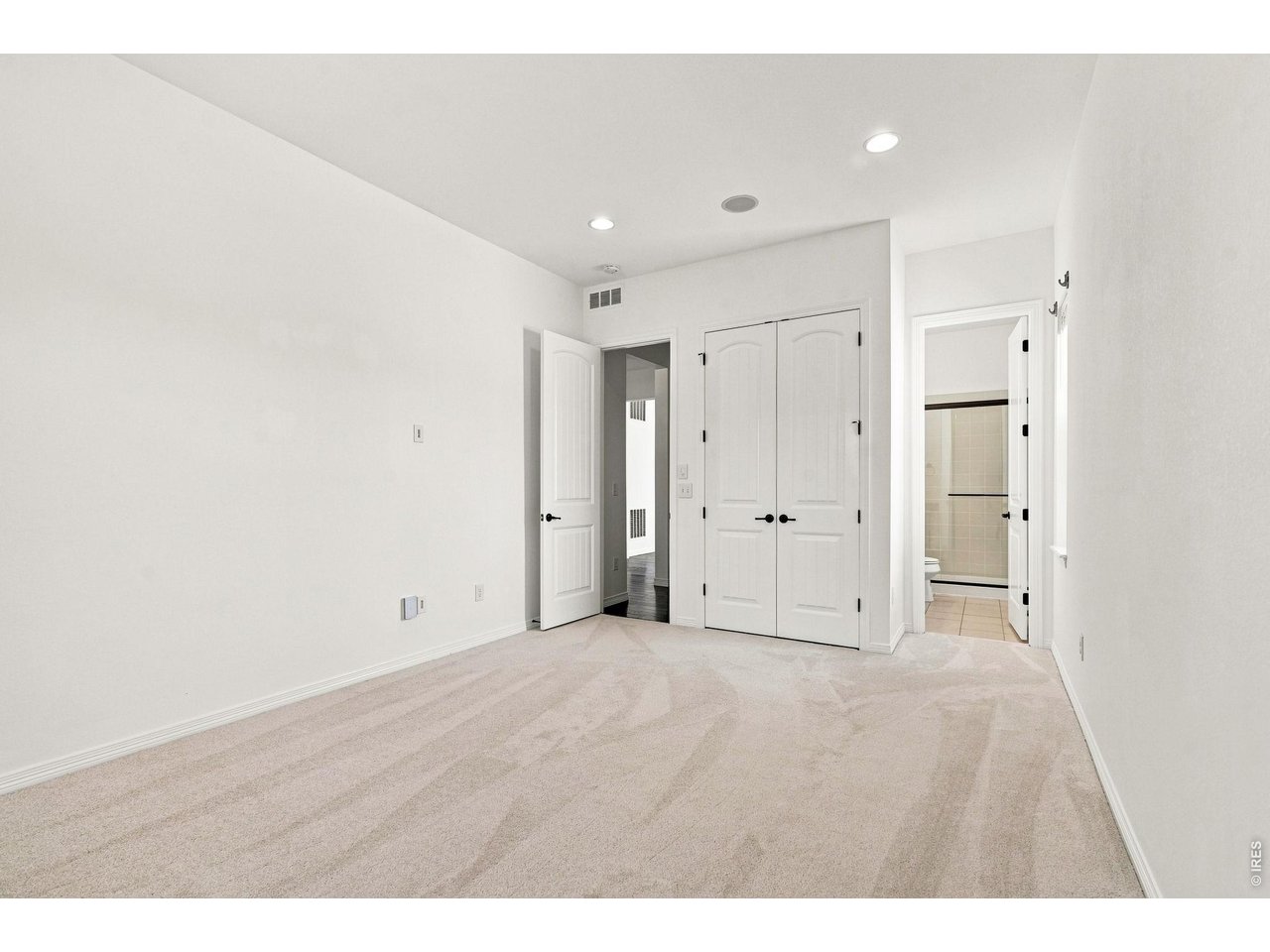 Empty bedroom with beige carpet, double closet doors, and an open doorway to a tiled bathroom.
