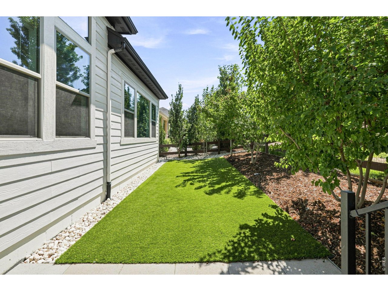 Side yard with a green lawn, white stone edging, and mature trees beside a light-colored home.