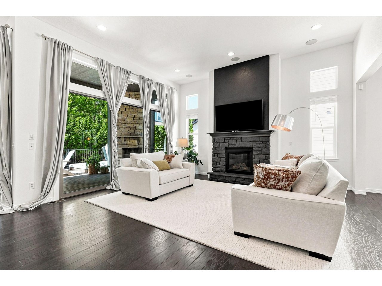 Staged living room with white sofas, a dark stone fireplace, and tall windows opening to a deck.