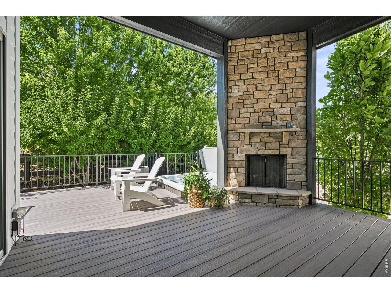 Covered deck with a stone fireplace, lounge chairs, and leafy trees beyond the railing.